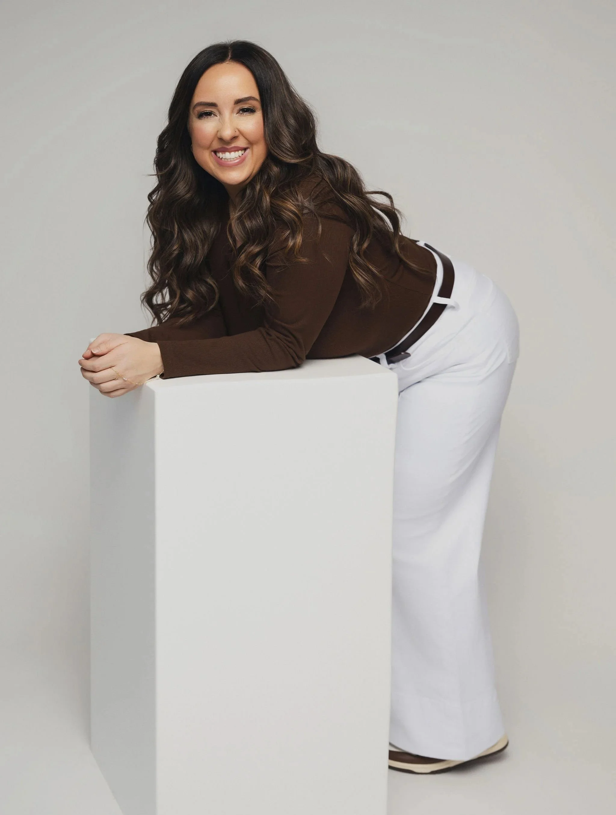 Woman with long, wavy brown hair leaning on a white rectangular block, smiling at the camera, wearing a brown top and white pants.