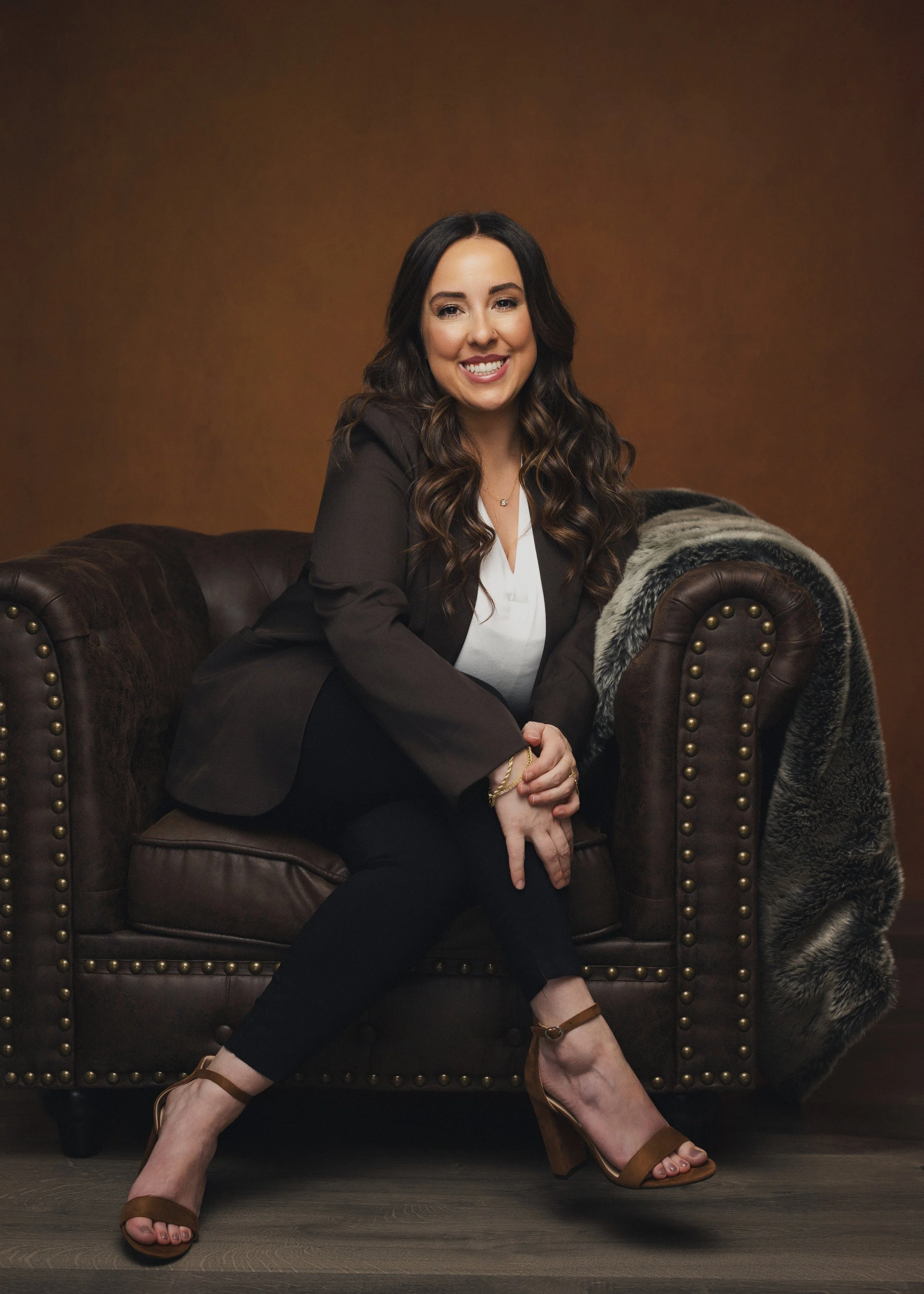 A woman with long wavy brown hair, dressed in a dark blazer, white blouse, and black pants, sitting on a brown leather sofa with a fur throw draped over the armrest, smiling at the camera. The background is a warm brown color.