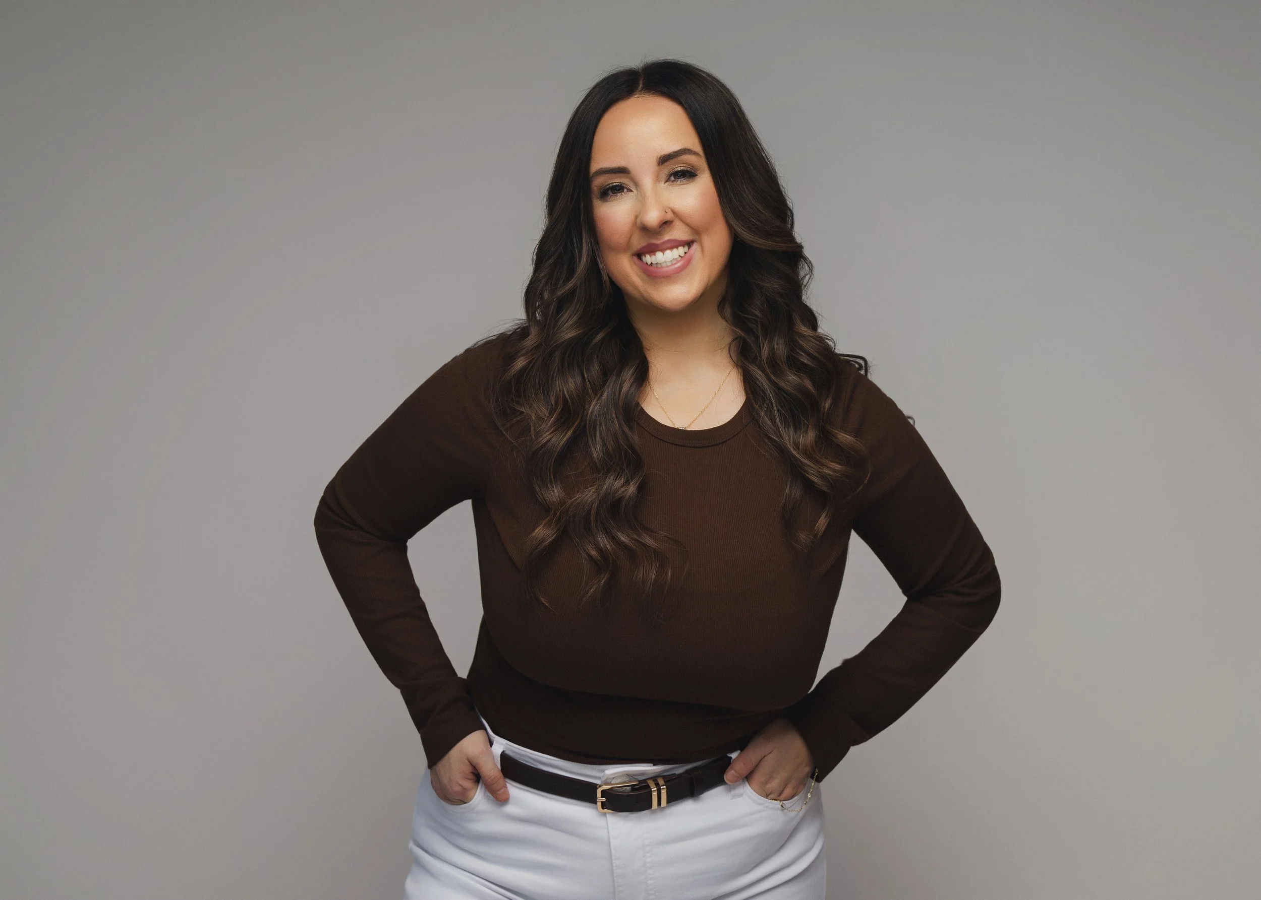A woman with long wavy brown hair smiling, wearing a brown long-sleeve shirt and white pants, standing with hands on hips against a gray background.