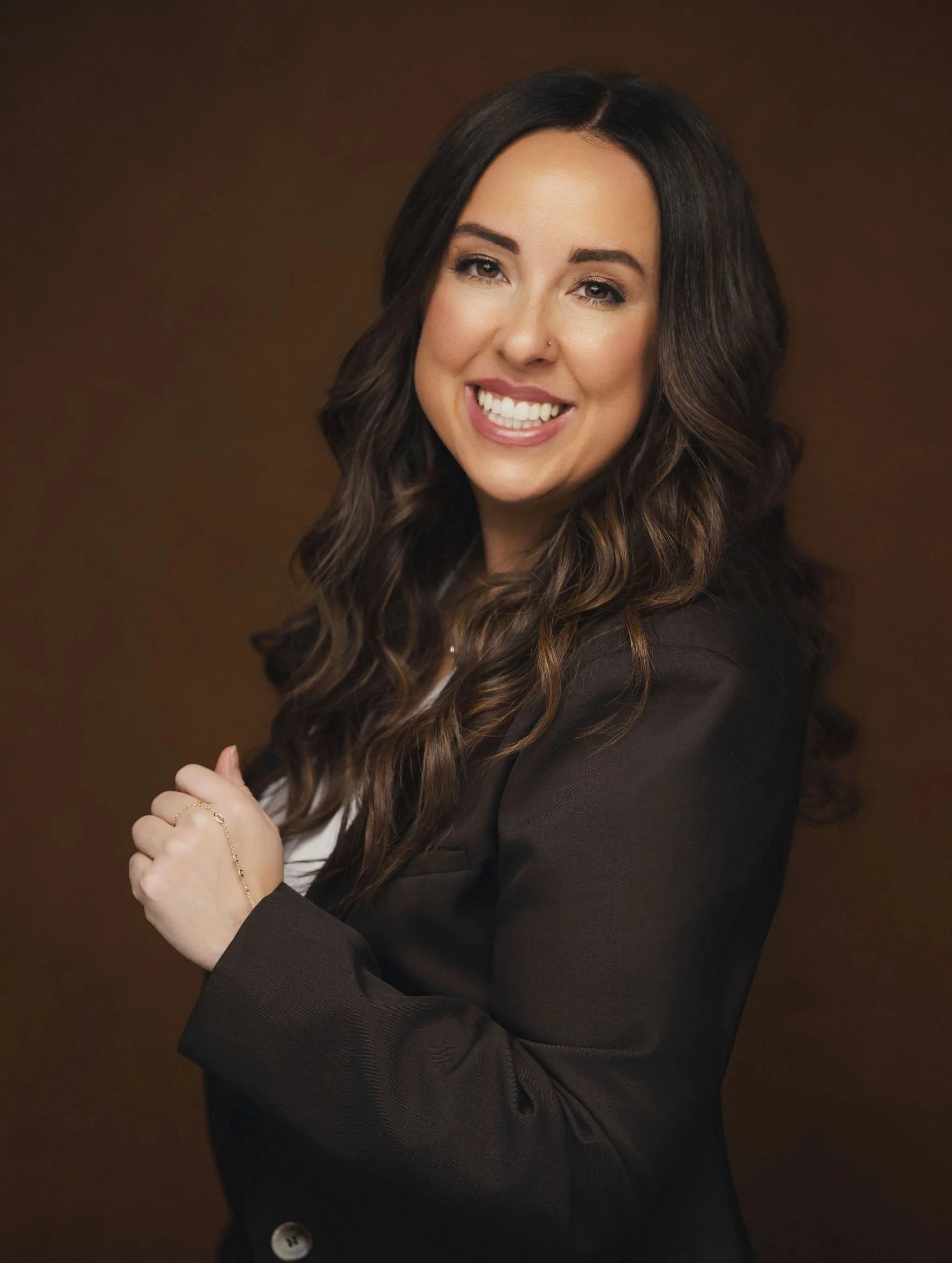 A woman with long, wavy brown hair wearing a black blazer, smiling, in front of a brown background.