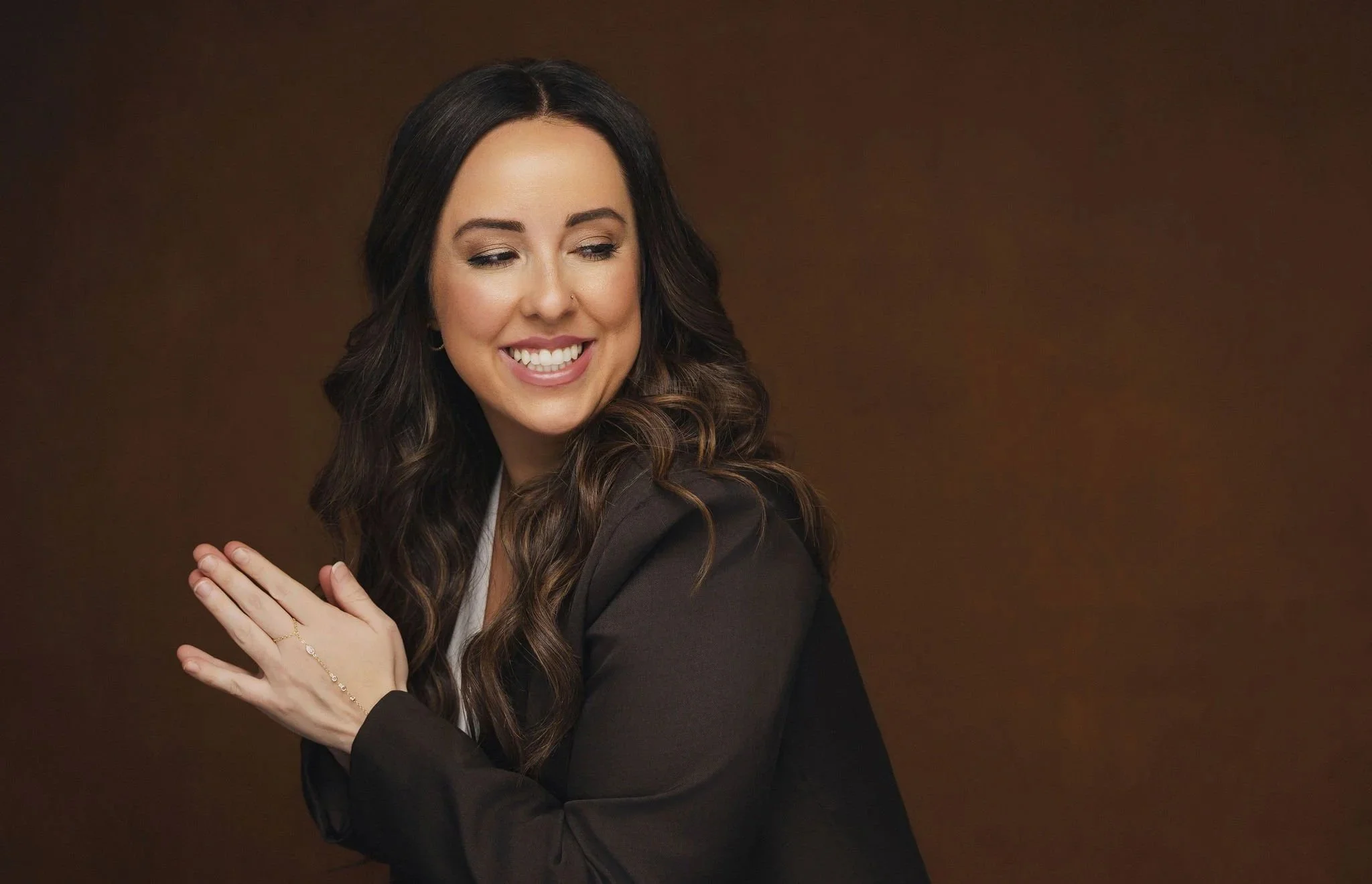 A woman with long dark brown hair smiling and looking to the side, wearing a black blazer and a white top, with one hand resting on her shoulder, against a brown background.