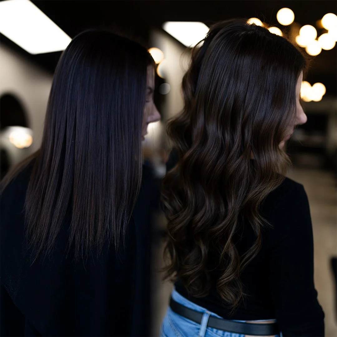 Two women with styled dark brown hair, one with straight hair and the other with wavy hair, in a salon setting with blurred lights in the background.