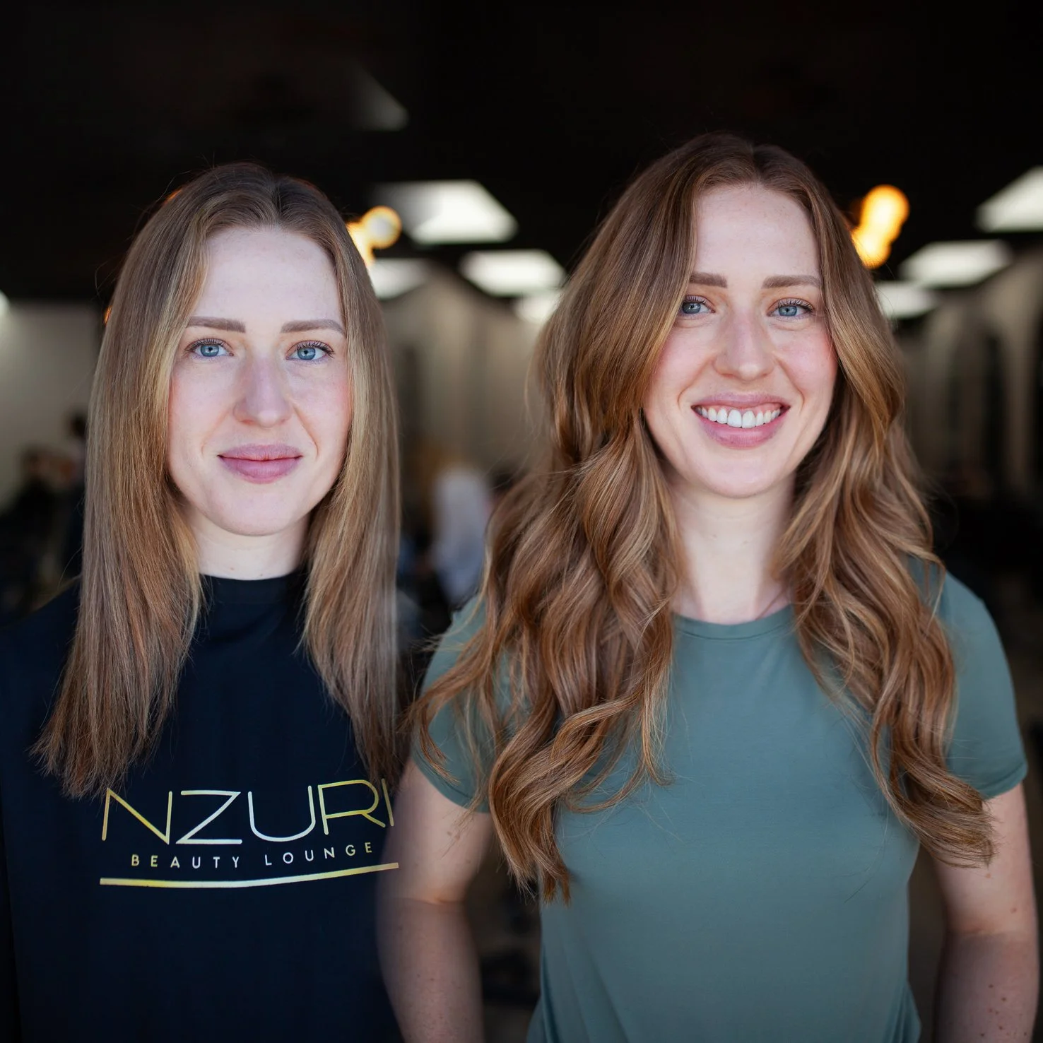Two women with long, wavy hair posing indoors, one smiling broadly and the other with a subtle smile, in a well-lit room with ceiling lights.