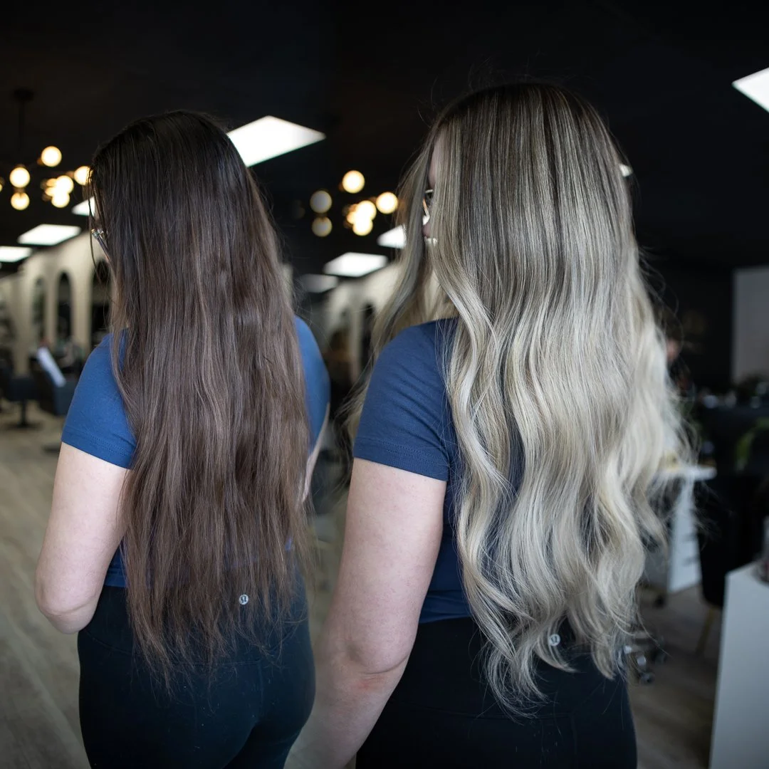 Two women with long wavy hair, one with dark blonde hair and one with platinum blonde hair, are standing in a hair salon.