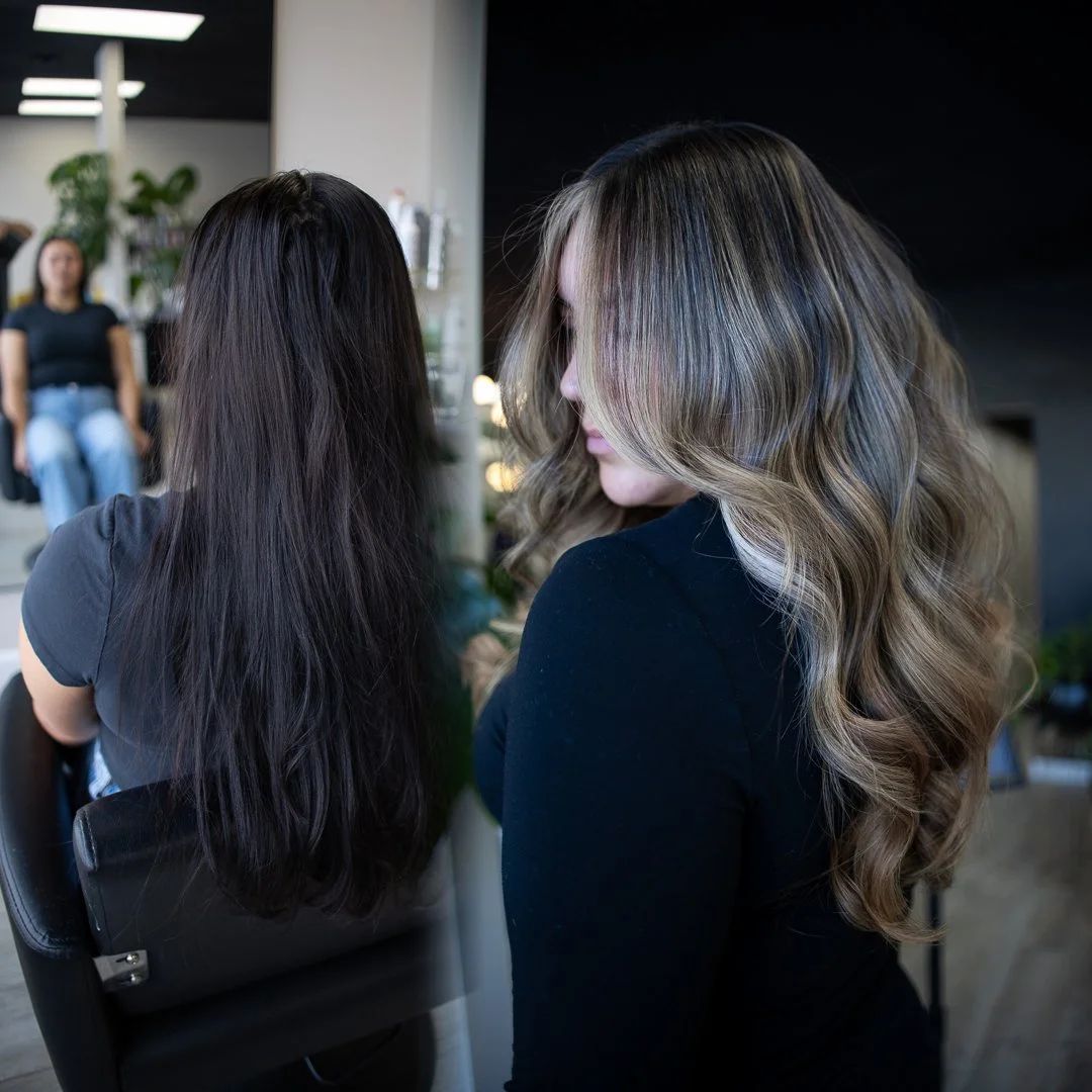 A woman with long wavy blonde hair looking at a woman with long straight dark hair in a salon.