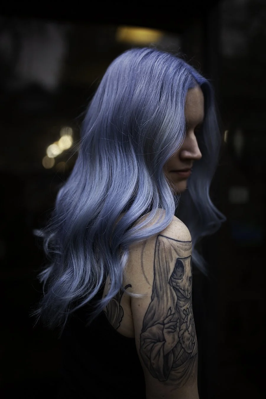 A woman with long wavy blue hair looking downward, showing a tattoo of a religious figure on her arm, in a dimly lit environment.