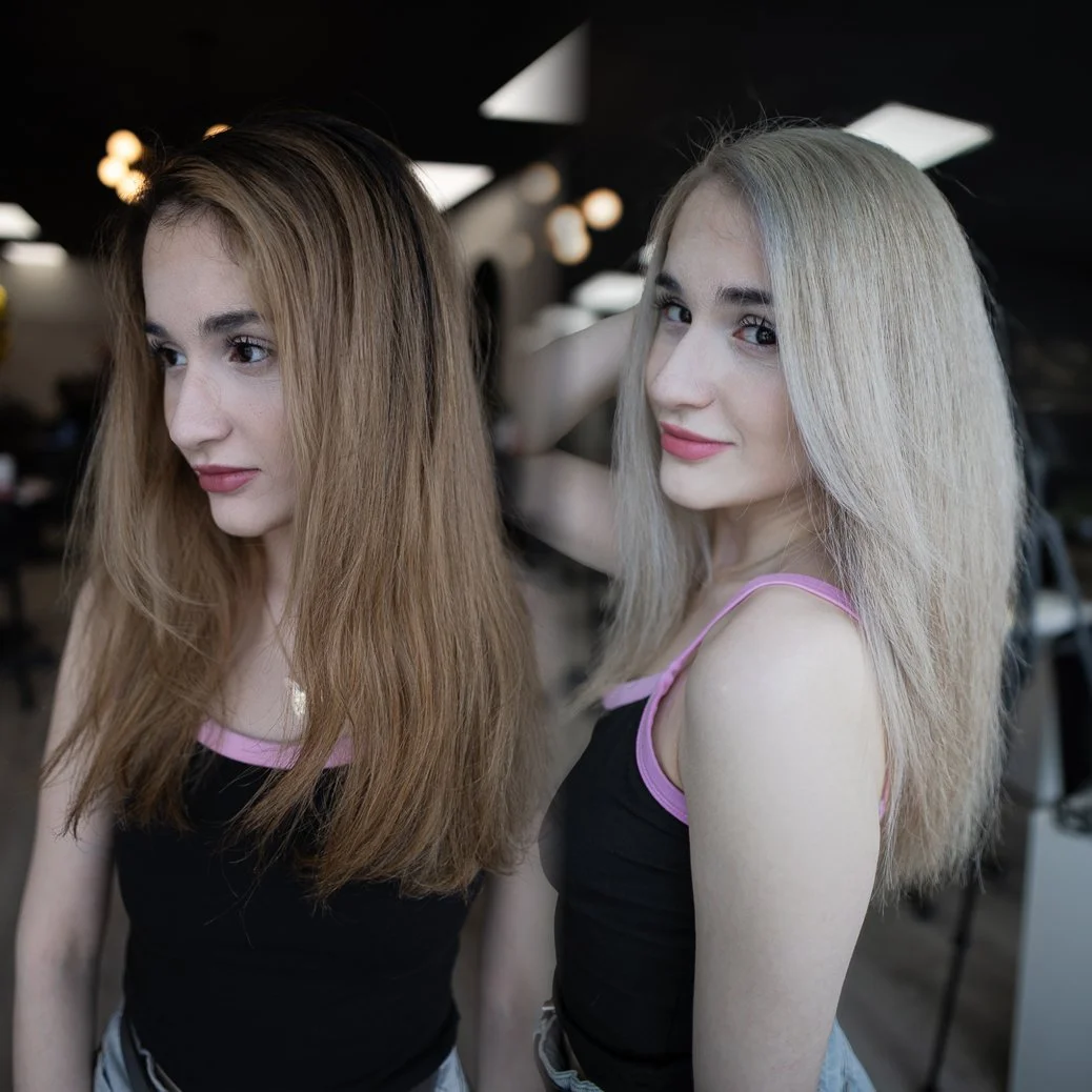 Two young women with styled hair posing in a salon or studio, one with brown hair and the other with blonde hair, both wearing black tops with pink straps.
