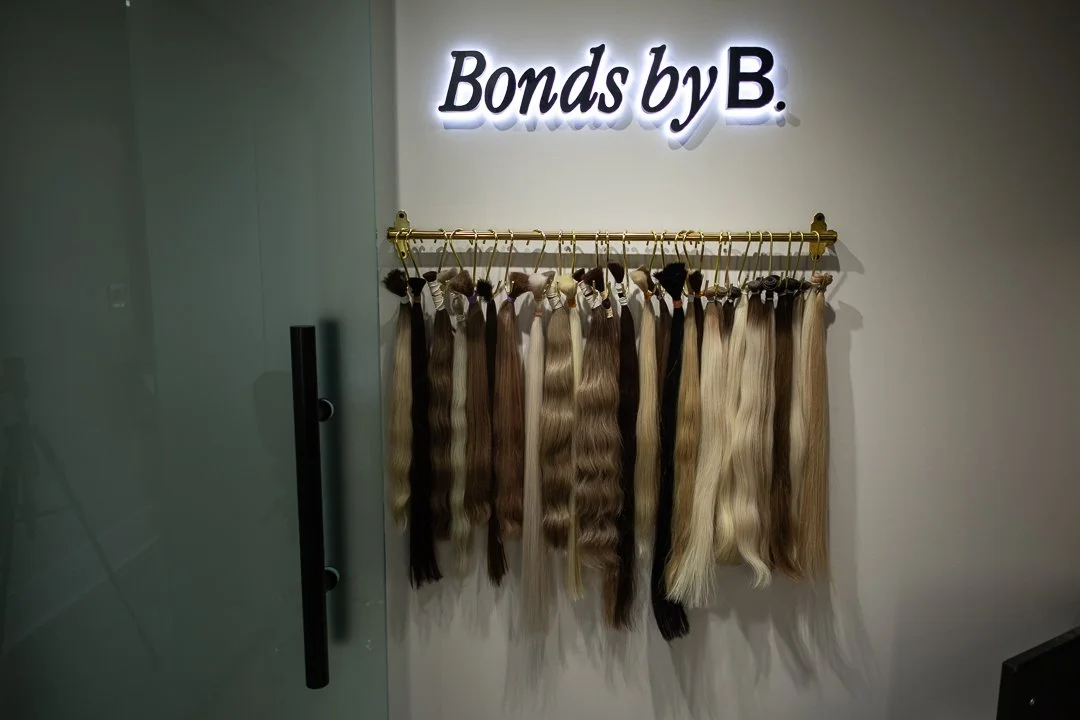Display of various hair extensions hanging on a gold rod in front of a white wall with a lit sign that reads "Bonds by B."