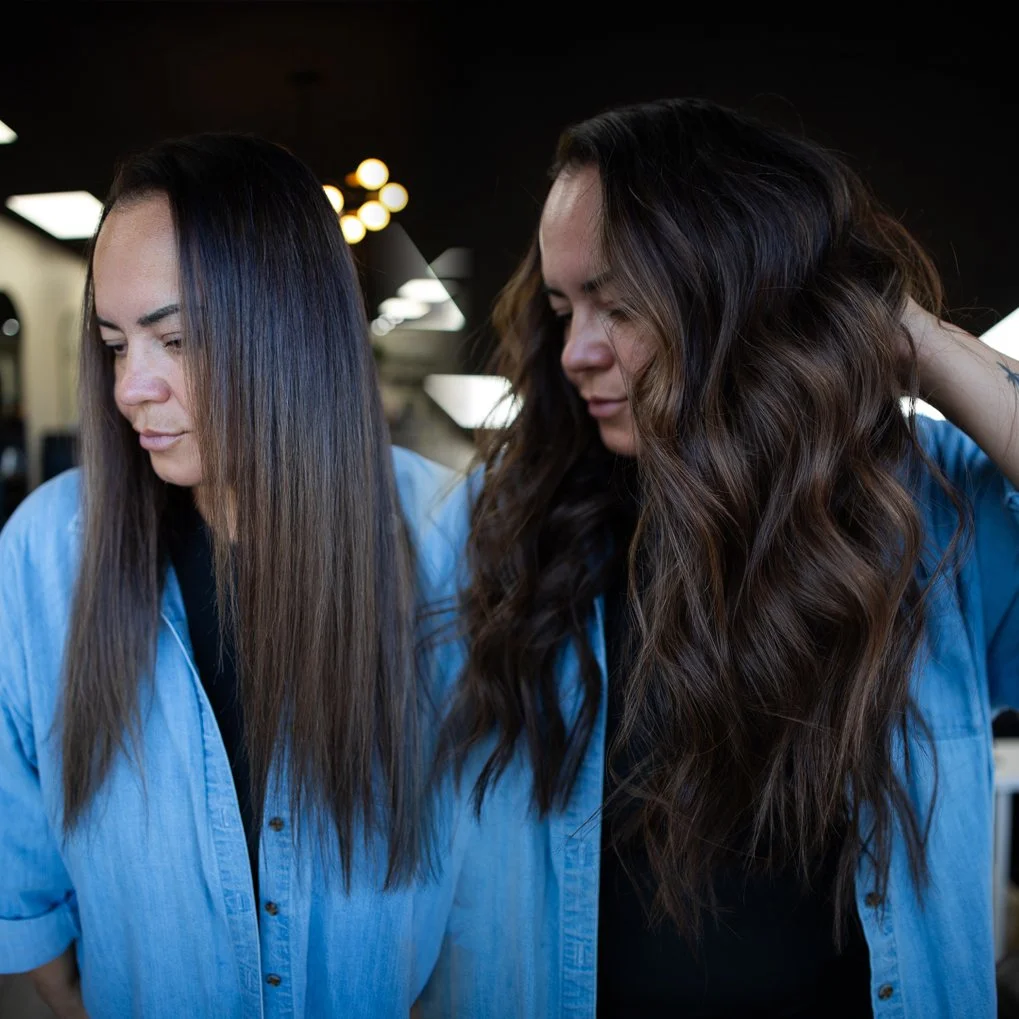 Two women with long hair, one with straight hair and the other with wavy hair, wearing blue shirts in a salon, looking down.
