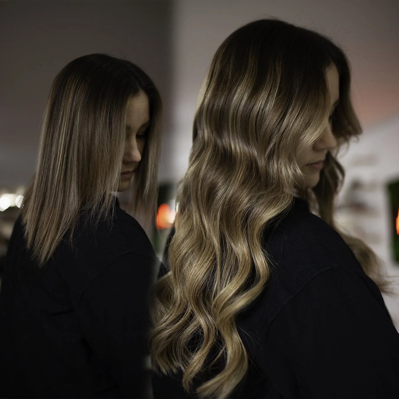 A woman with wavy blonde hair stands with her eyes closed, wearing a black shirt, in a dimly lit room with a second woman with straight, shoulder-length blonde hair close behind her.