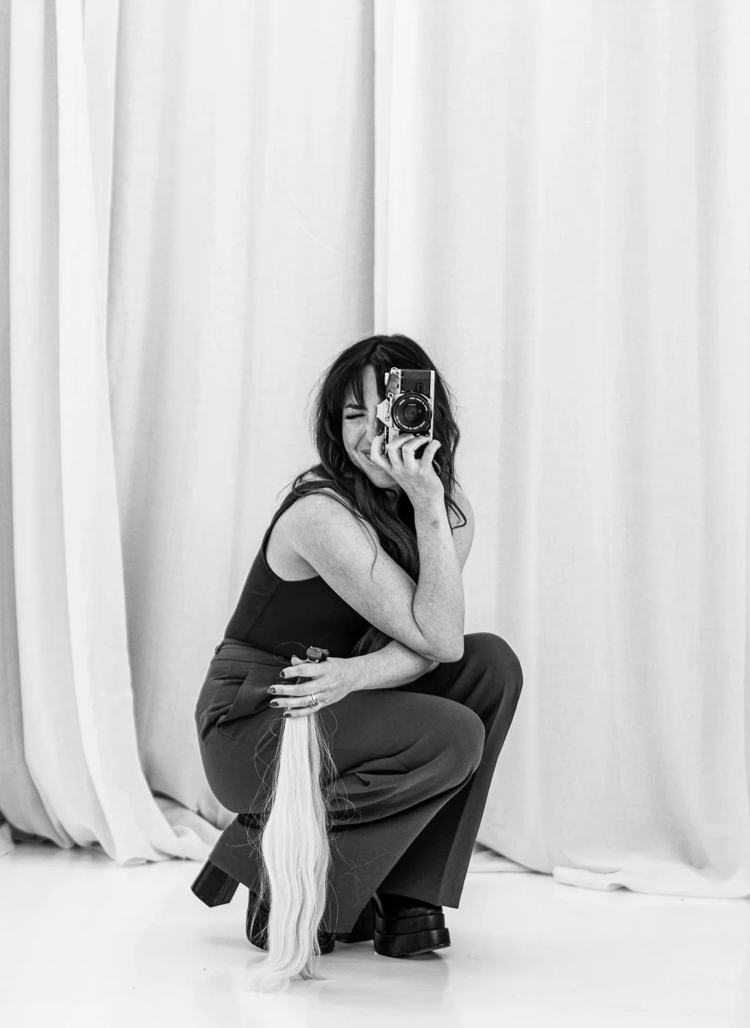 Woman with dark hair in an outfit squatting, holding a camera up to face, taking a picture in front of curtains.