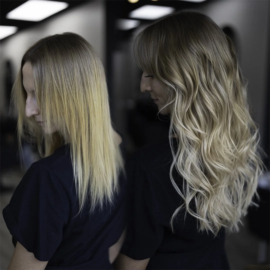 Two women with styled blonde hair in a salon, side profiles, one with loose wavy hair and the other with slightly tousled straight hair, wearing black shirts.