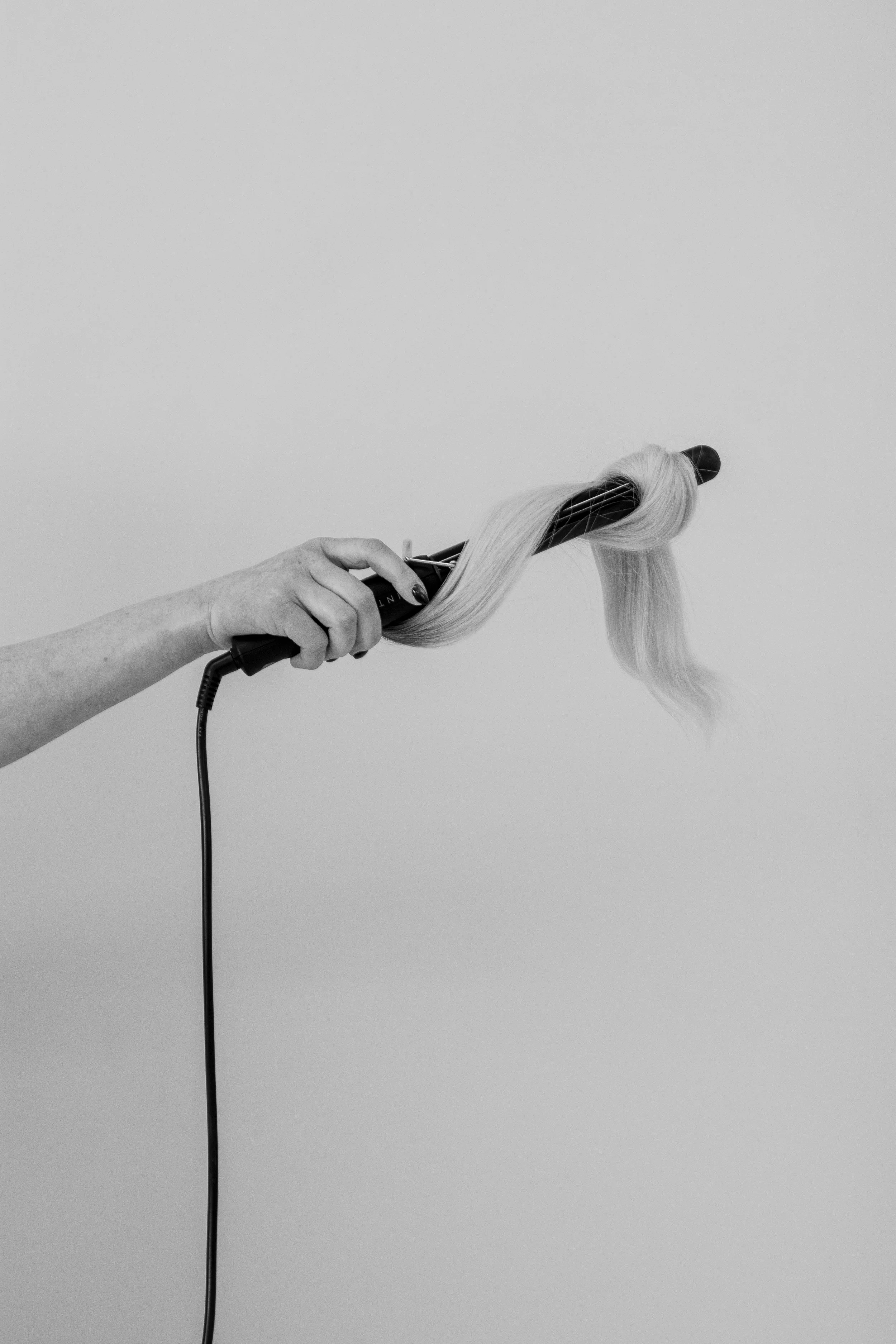 Hand holding a curling iron with blonde hair wrapped around it against a plain background.