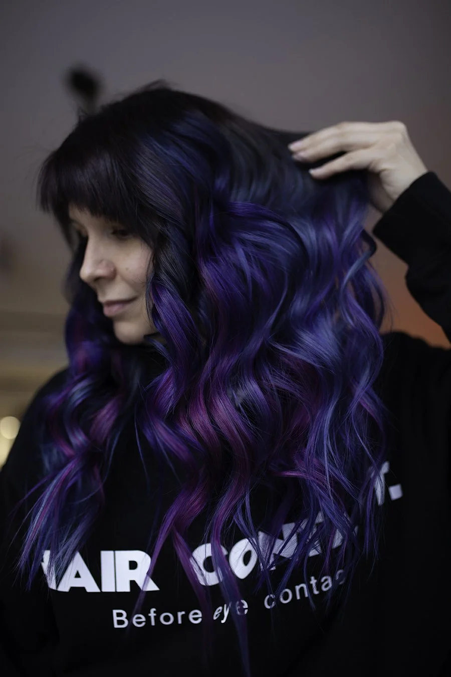 Woman with long wavy hair dyed in black, blue, purple, and pink, touching her hair with her right hand, wearing a black sweatshirt with white text.