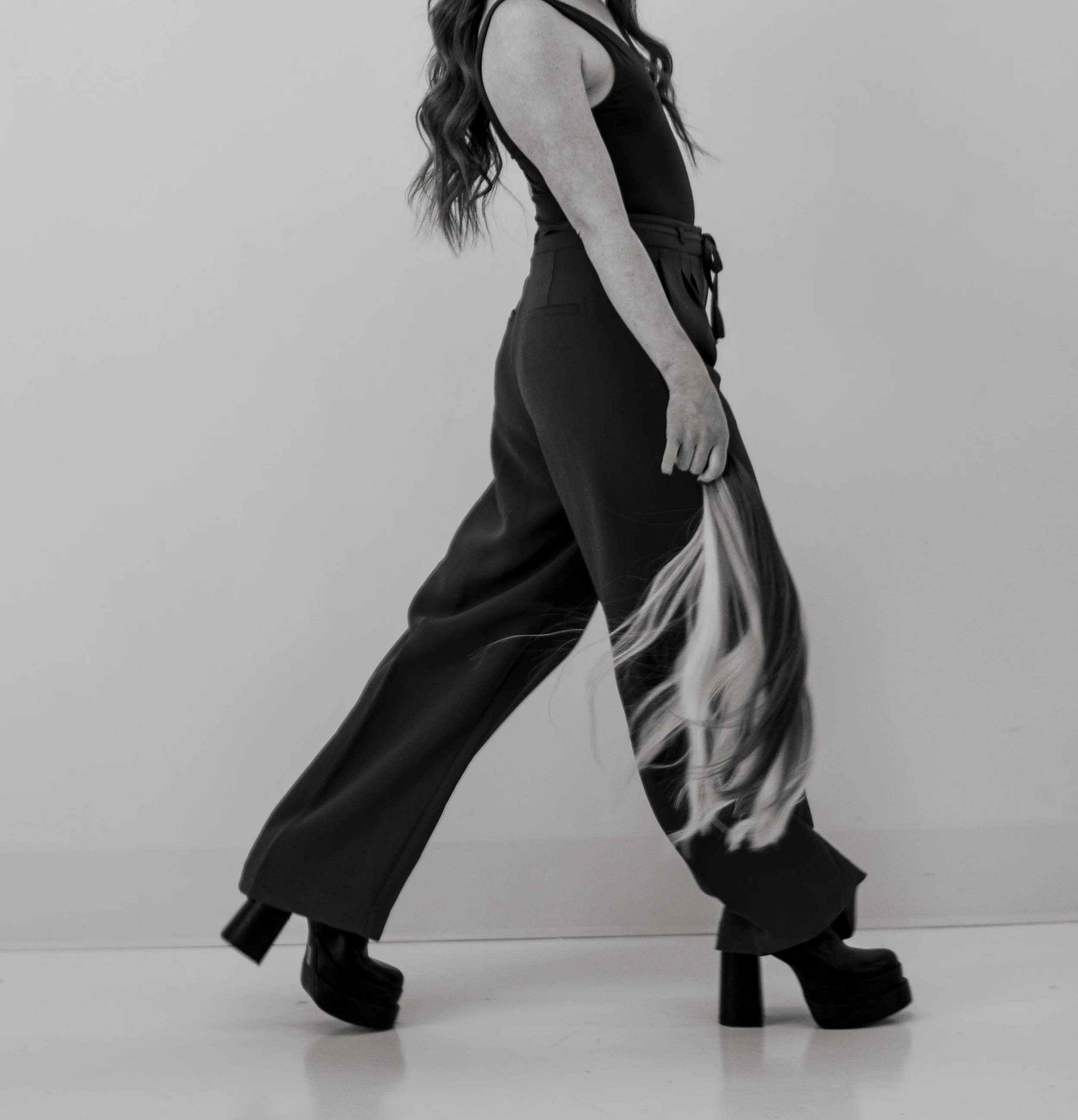 A woman with long hair walking in black platform shoes, wide-leg pants, and a sleeveless top, holding a long piece of fabric, in a minimal indoor setting.