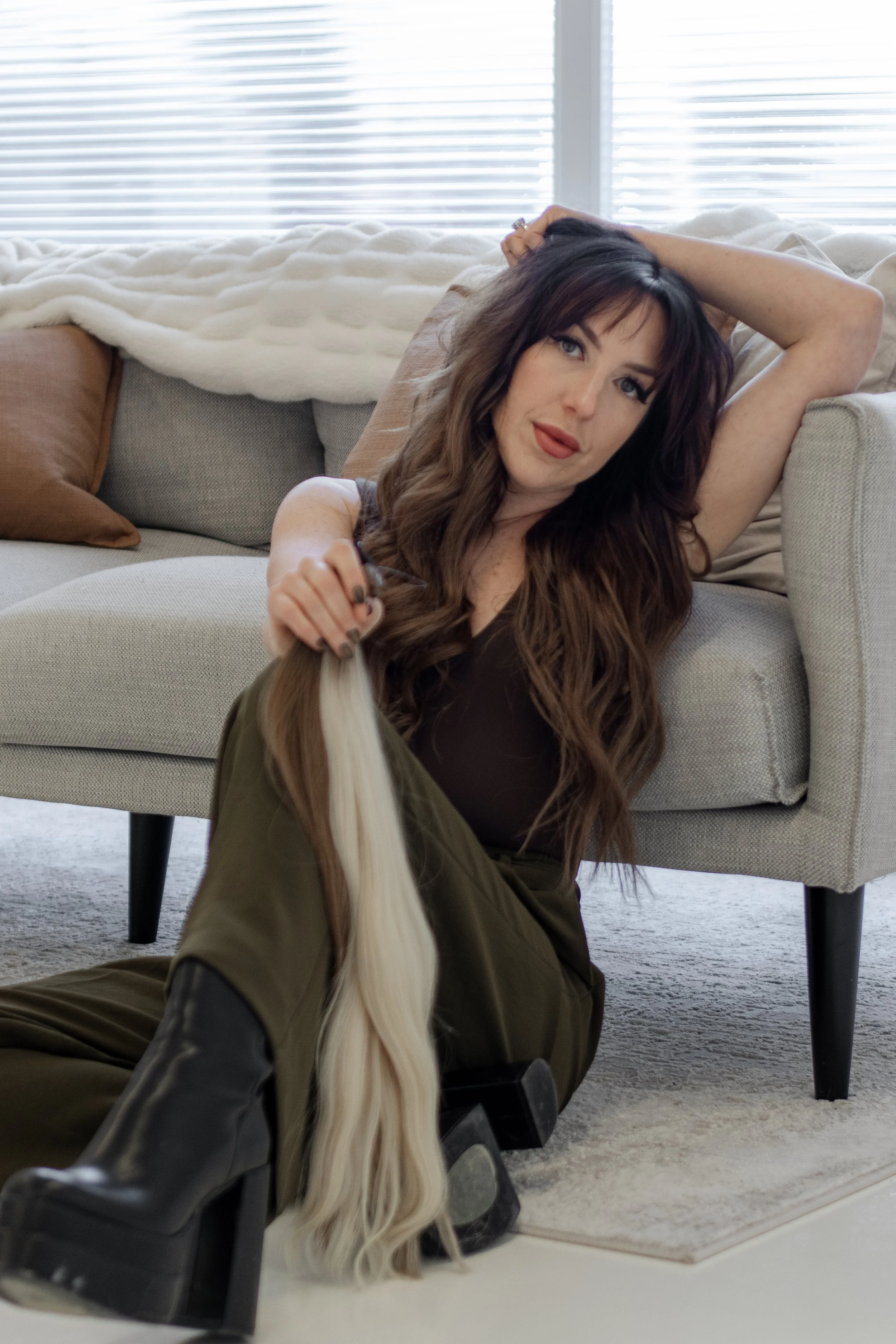 A woman with long brown hair, holding a blonde hair extension, sitting on the floor next to a beige sofa with brown and beige pillows, in a living room with white blinds.