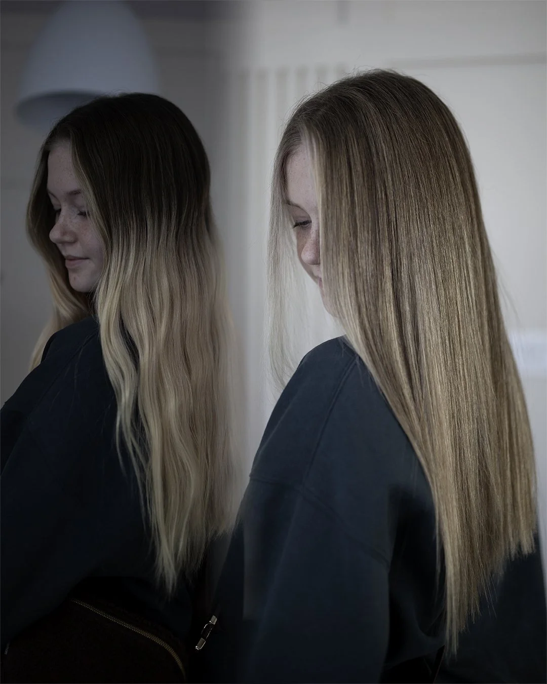 Side view of a girl with long, straight, reddish-blonde hair, smiling and looking down, standing beside a mirror showing her reflection.