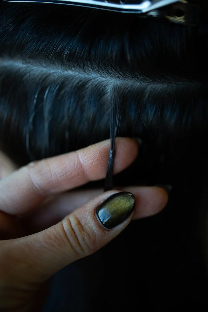 A hand holding a strand of black hair near the scalp, with the person's nails painted black. The hair appears to be being examined or styled.