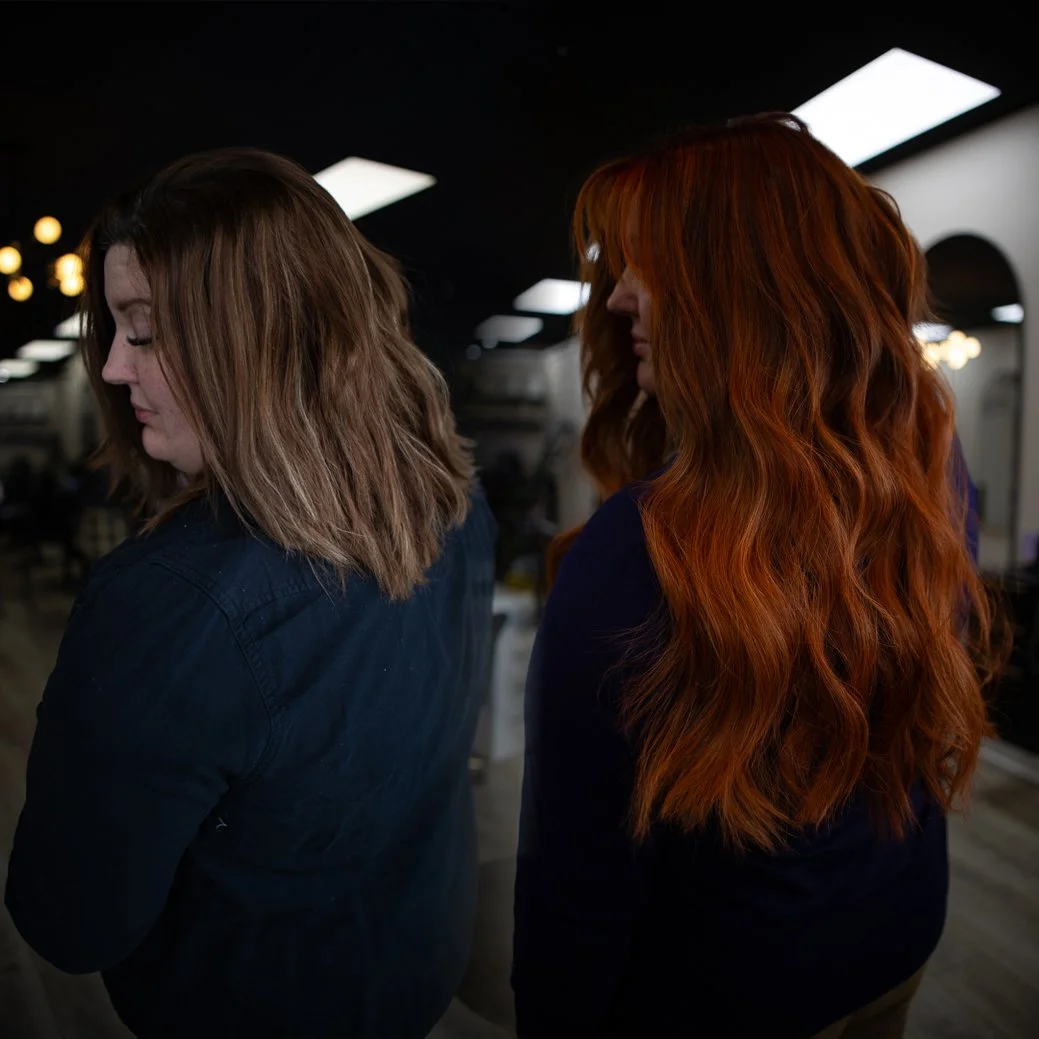 Two women with shoulder-length hair, one with light brown hair and the other with long, red, curly hair, standing indoors in a room with black ceiling and bright ceiling lights.