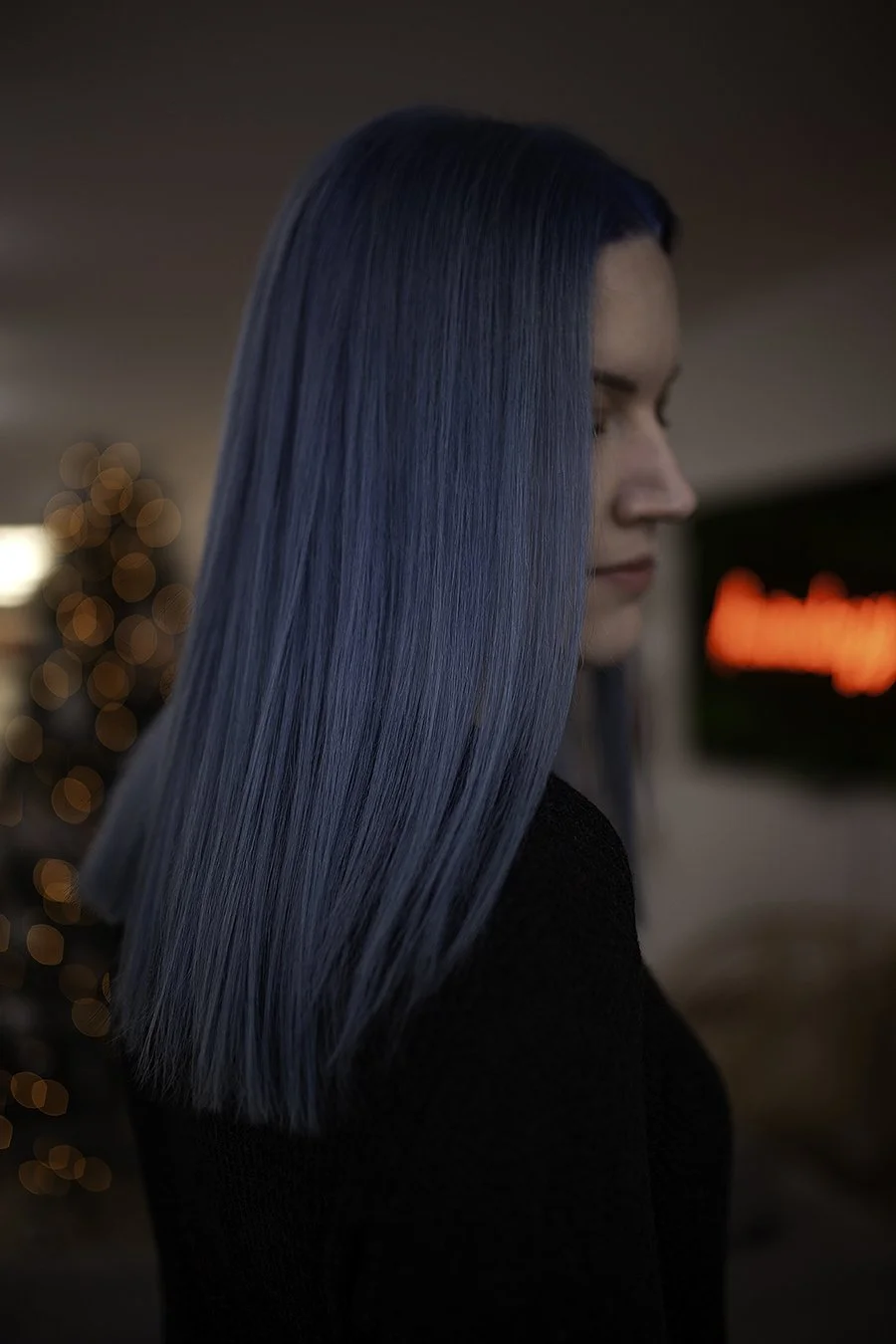 Profile of a woman with straight, vibrant blue hair against blurred warm ambient background.