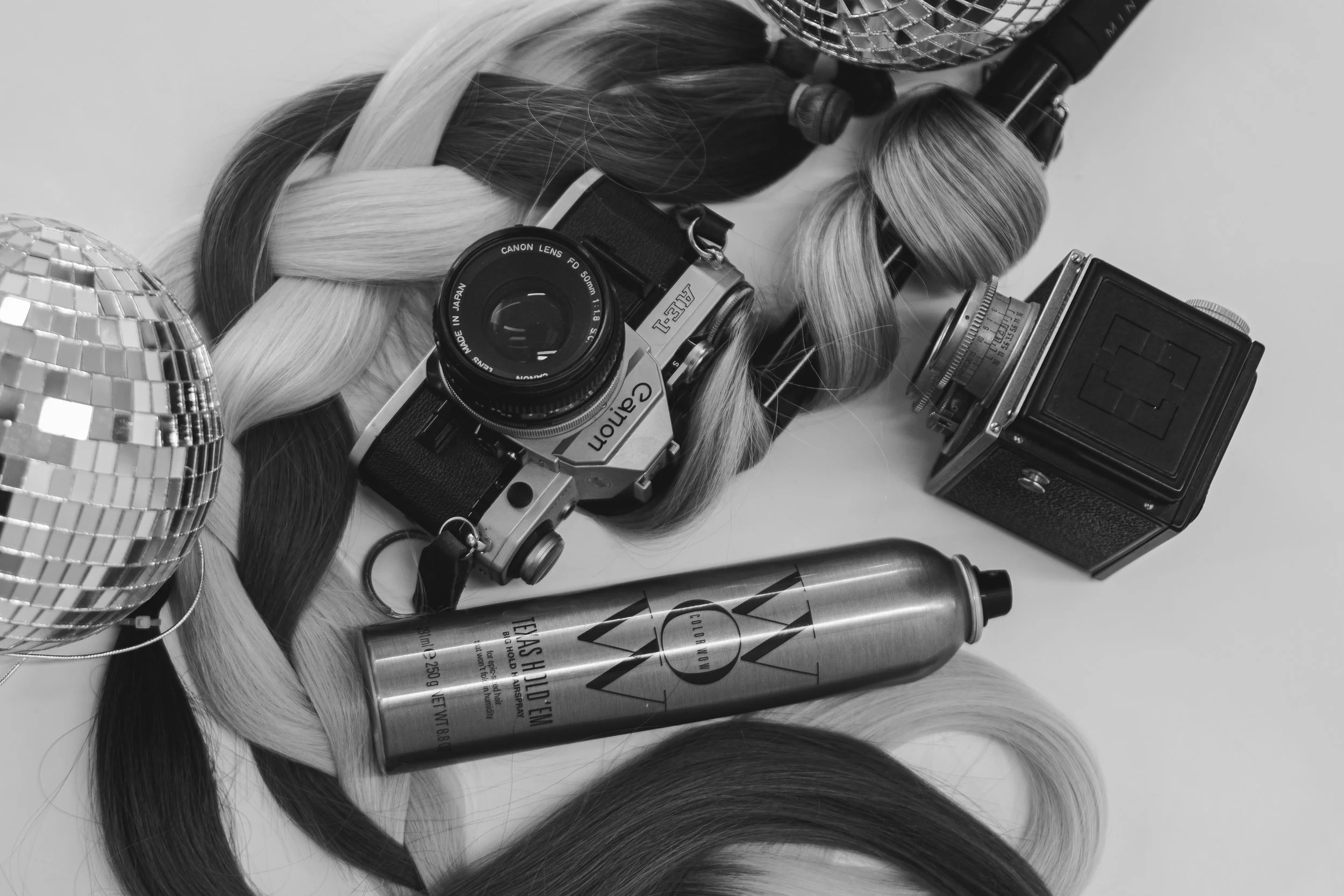 A black and white photo of hair styling tools, a vintage camera, a bottle of dry shampoo, and two disco balls laid on a surface.