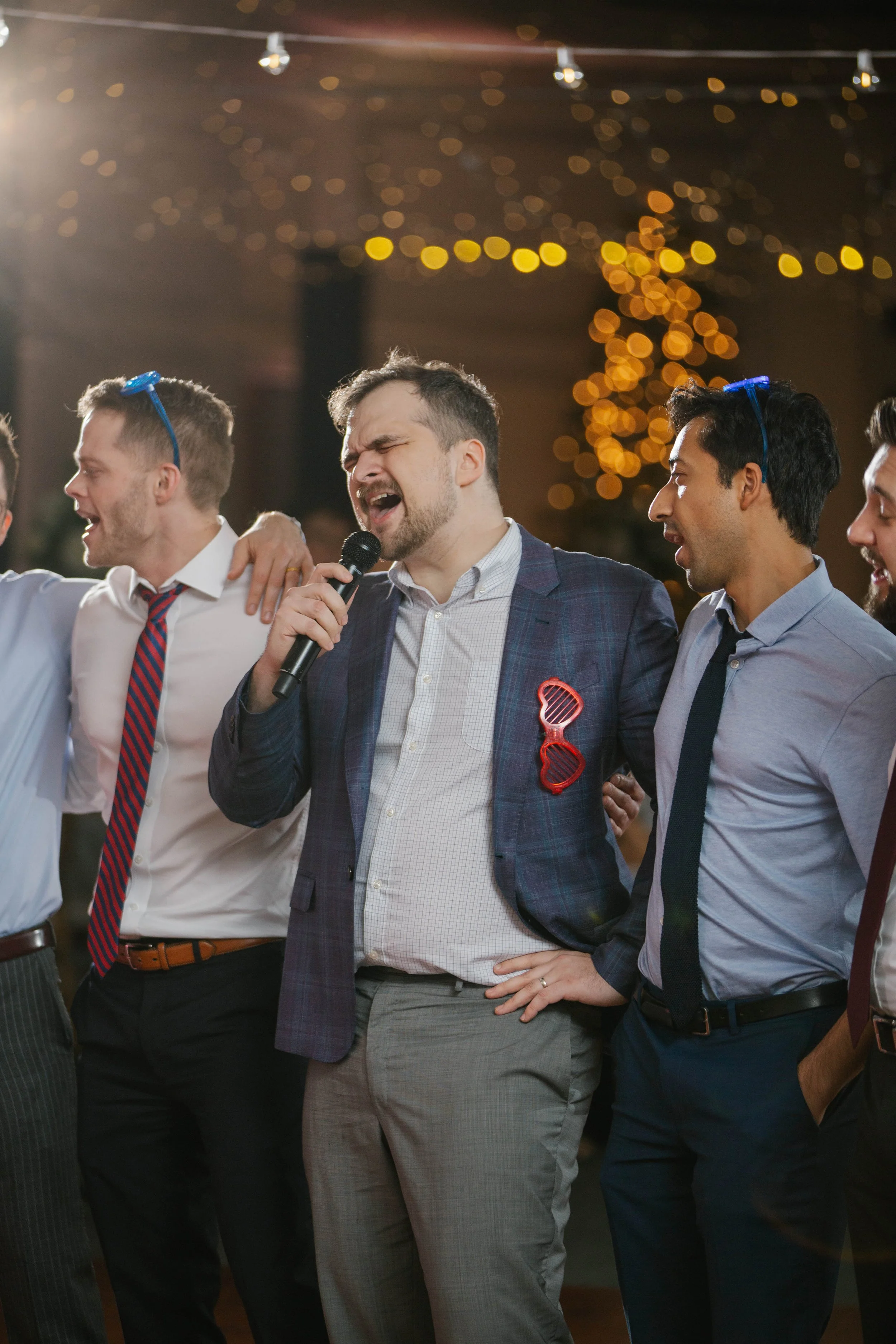 Group of men celebrating, with one man singing into a microphone, at a party or event with string lights in the background.