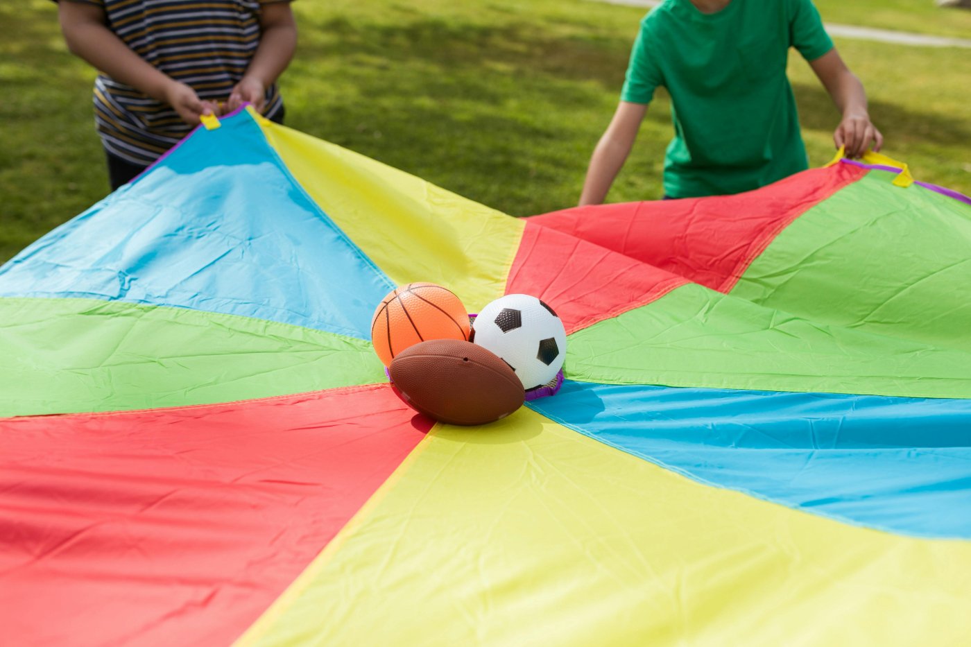 Children playing with a multicolored parachute outdoors, with sports balls including a basketball, soccer ball, and football on the parachute.