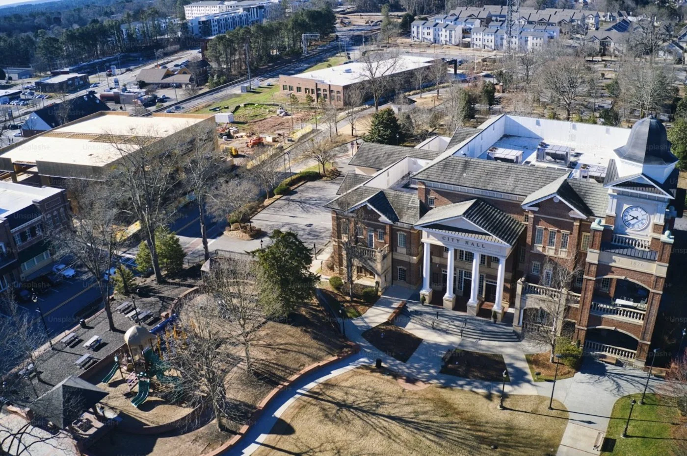 Aerial view of a city hall building with a clock tower, surrounded by trees, parking lots, and various buildings in a mid-size downtown area