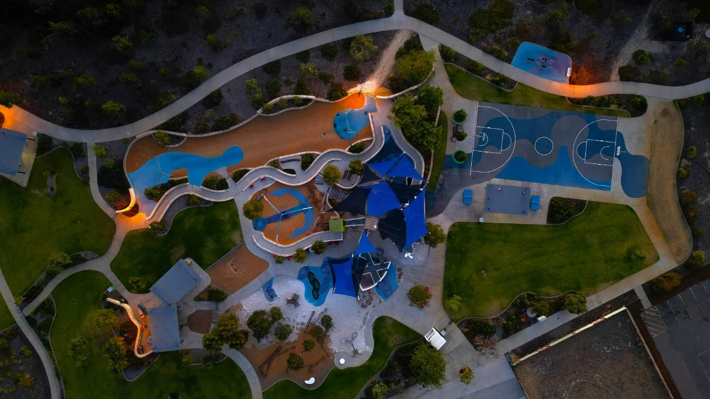 Aerial view of a colorful urban park featuring winding paths, playgrounds, a basketball court, and green lawns, illuminated with warm lighting during dusk.