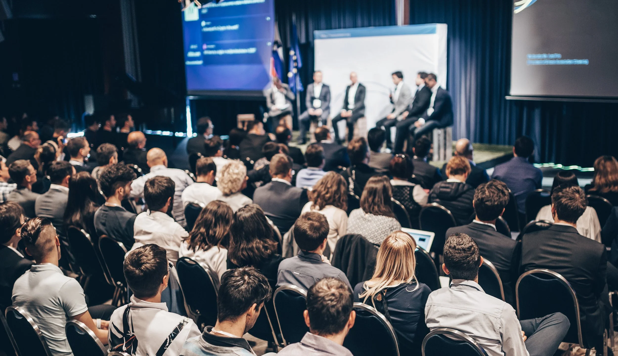 A large audience attending a professional conference with four speakers on stage in formal attire, seated and speaking into microphones, with presentation screens behind them.