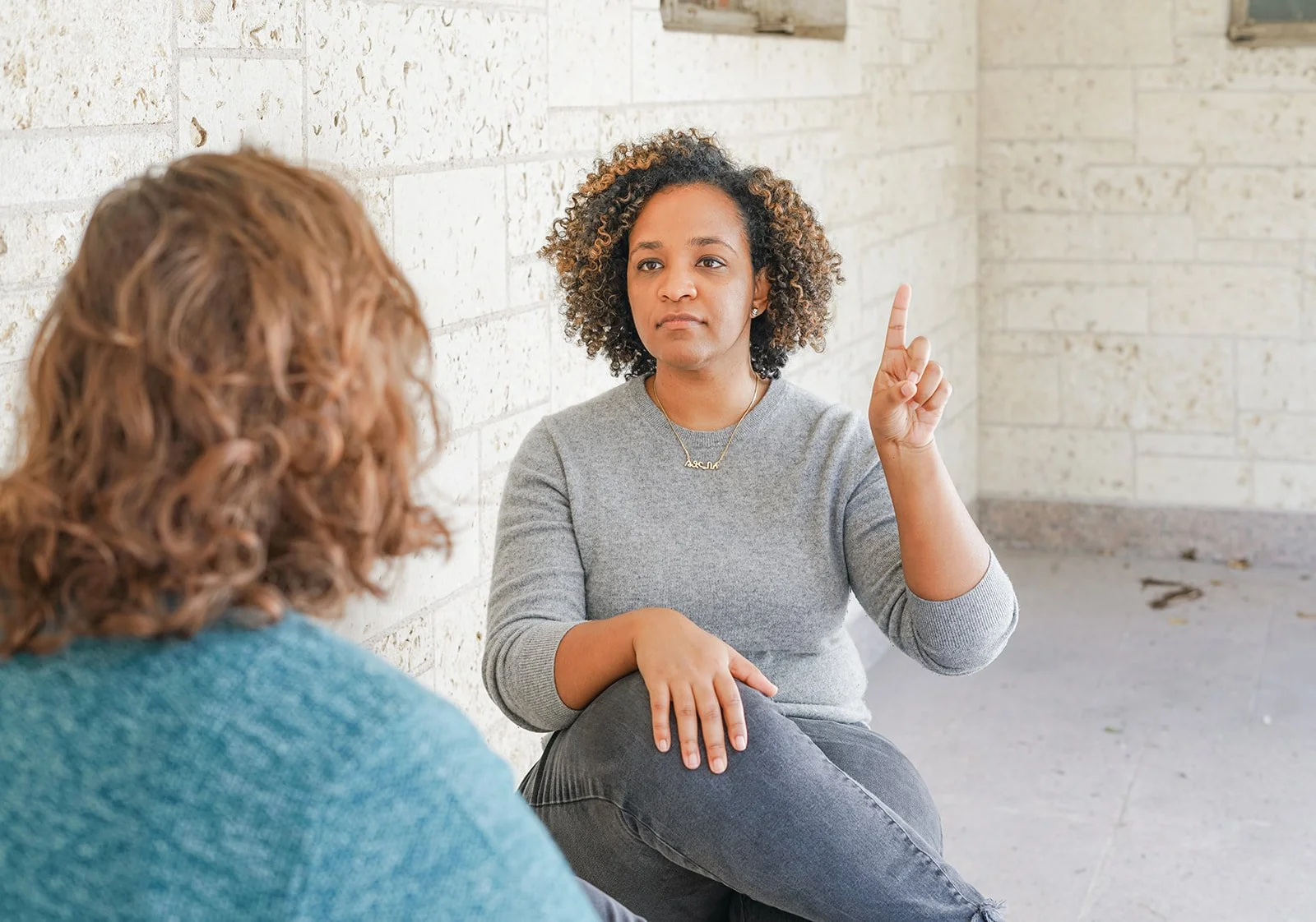 Two women having a conversation outdoors. One woman is pointing up with her finger, sitting against a brick wall. The other woman, with curly hair, listens to her.