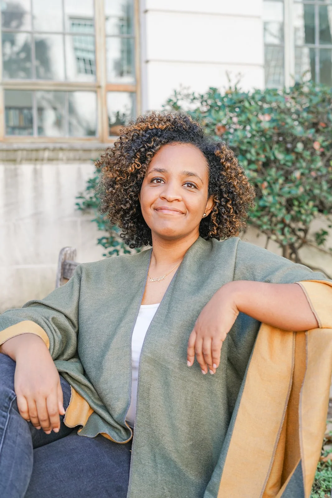 A woman with curly hair sitting outdoors on a wooden chair, smiling, in front of greenery and a building with glass windows.