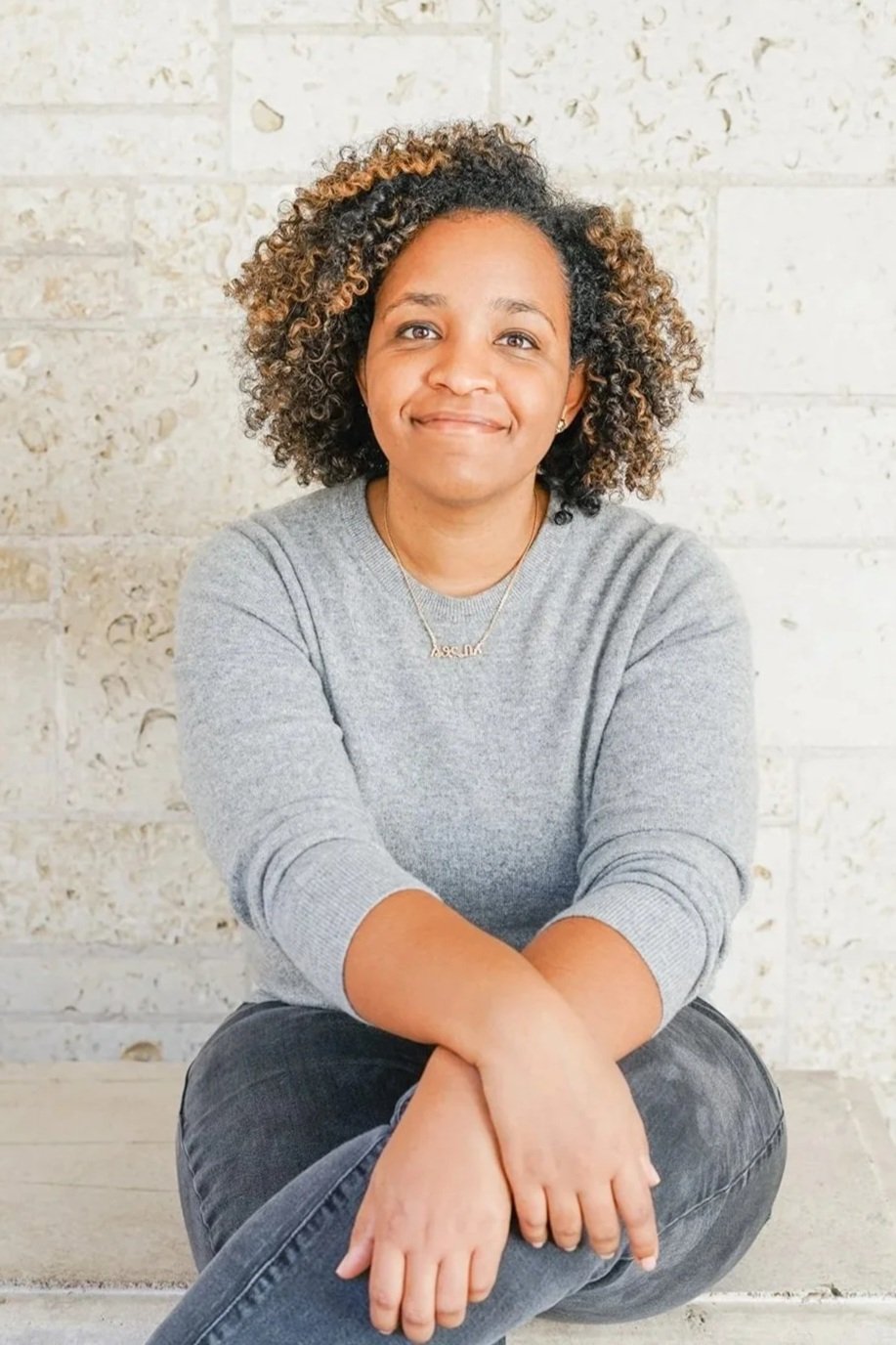 A woman with curly hair, wearing a gray sweater and black jeans, sitting on the ground with a smile against a light-colored brick wall.