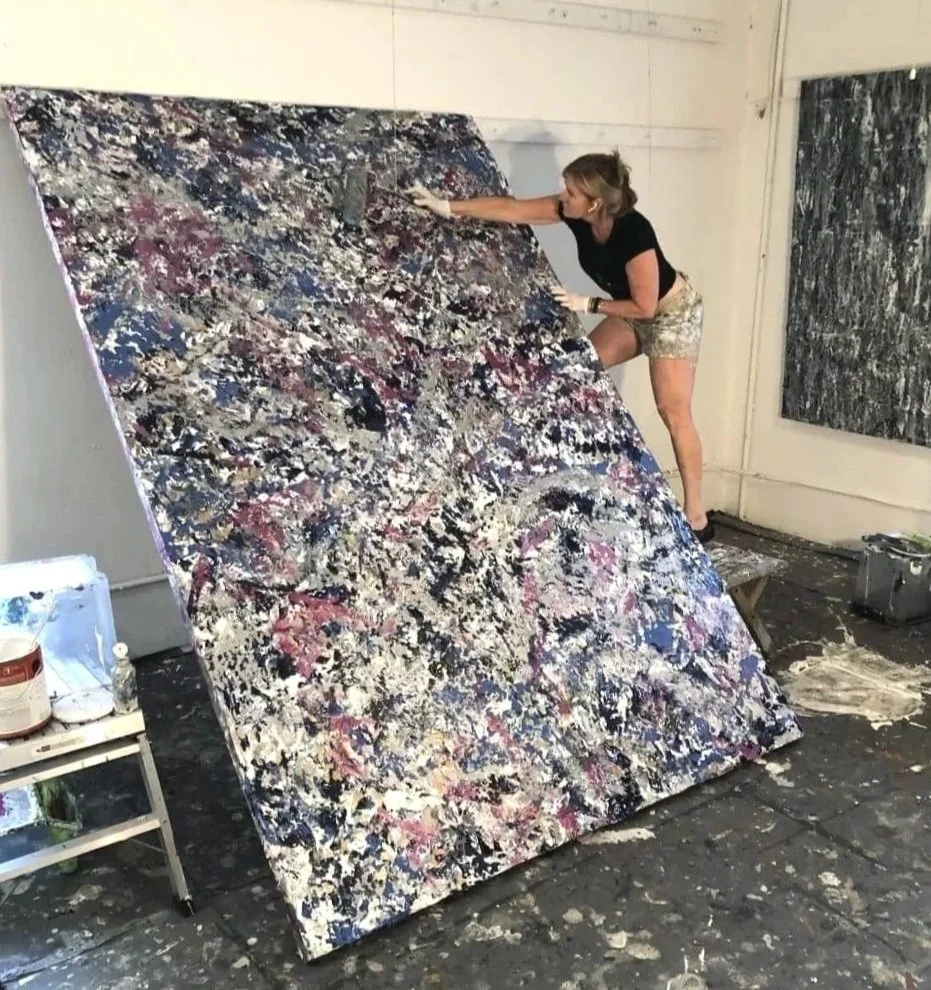An artist is working on a large abstract painting in a studio. The painting features a chaotic blend of colors including black, white, pink, and blue. The artist is standing on a bench, leaning over, and using a tool to create texture on the canvas.