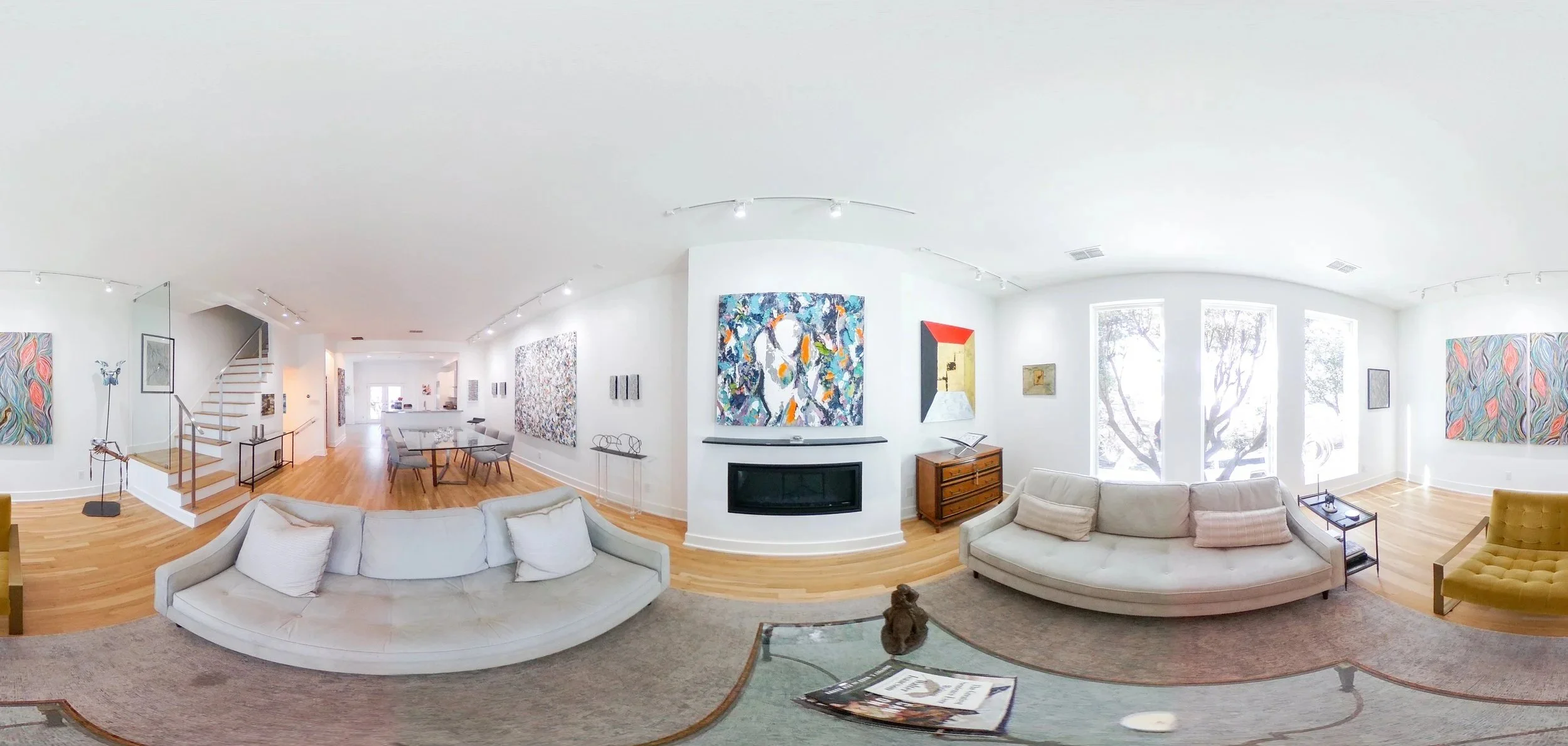 Spacious living room with white walls, hardwood floors, large abstract paintings, a white curved sofa, a yellow armchair, a glass coffee table with books and a teddy bear, and floor-to-ceiling windows with trees outside.