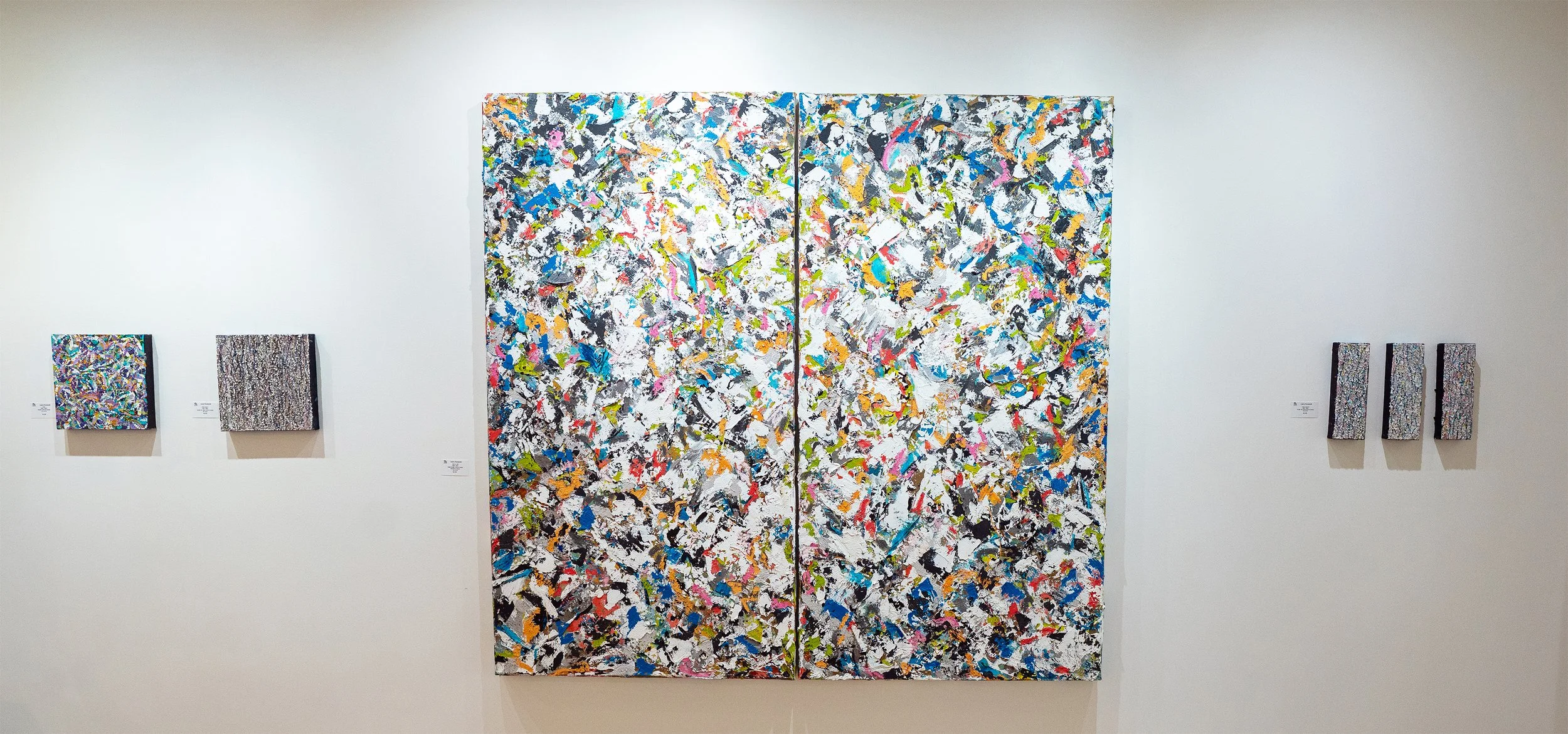 Gallery wall with five abstract paintings, including a large diptych center piece with multicolored splashes on a white background, and four smaller paintings with similar abstract patterns, all hung on a white gallery wall.