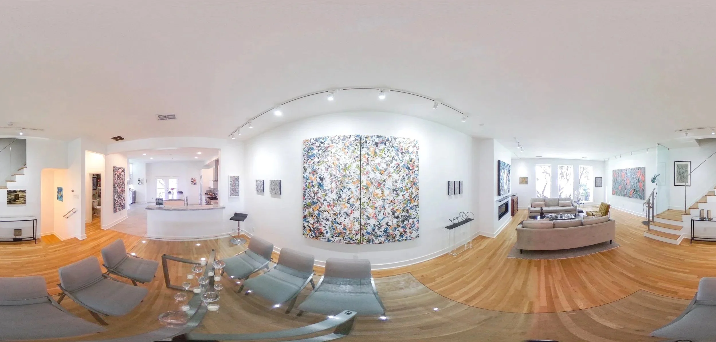 Panoramic view of a modern, well-lit art gallery with various abstract paintings on white walls, wooden flooring, a sitting area with sofas, and a staircase.