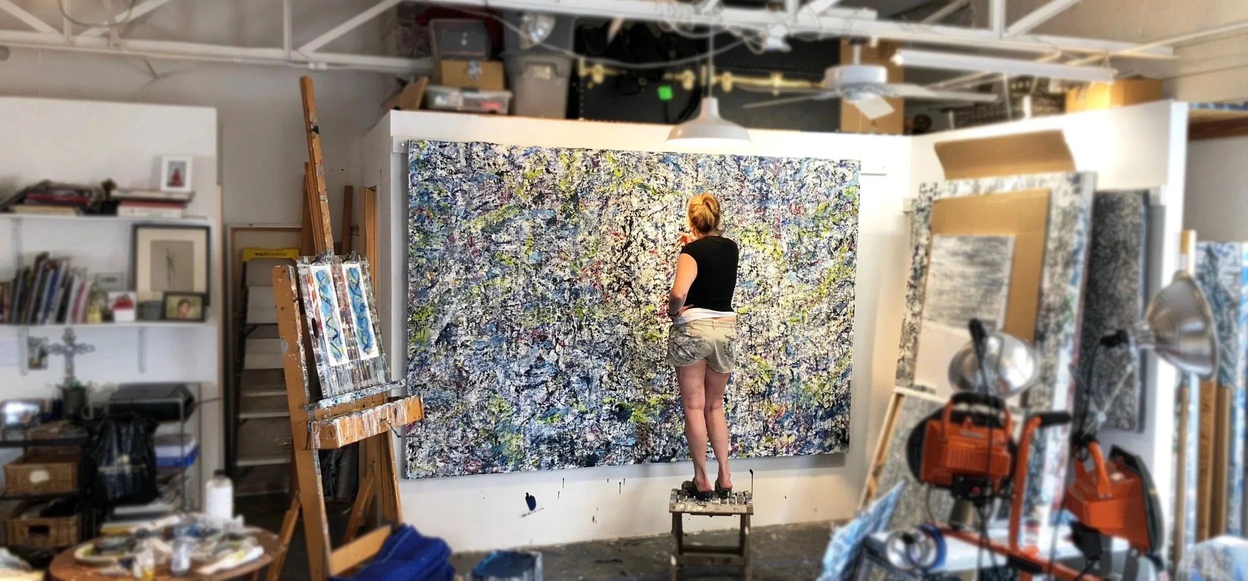 An artist standing on a small platform, working on a large abstract painting with splashes of various colors on a wall in an art studio.
