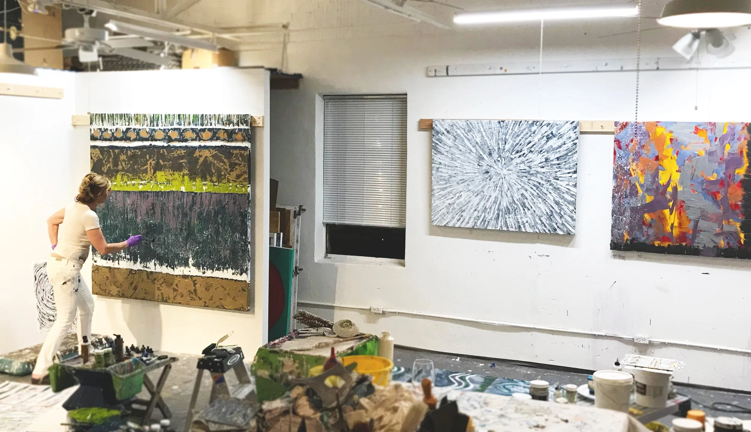 An artist in a white outfit and purple gloves is working on a large abstract painting in a studio. The studio has three other large abstract paintings on the wall, and various art supplies and tools are scattered on a table in the foreground.