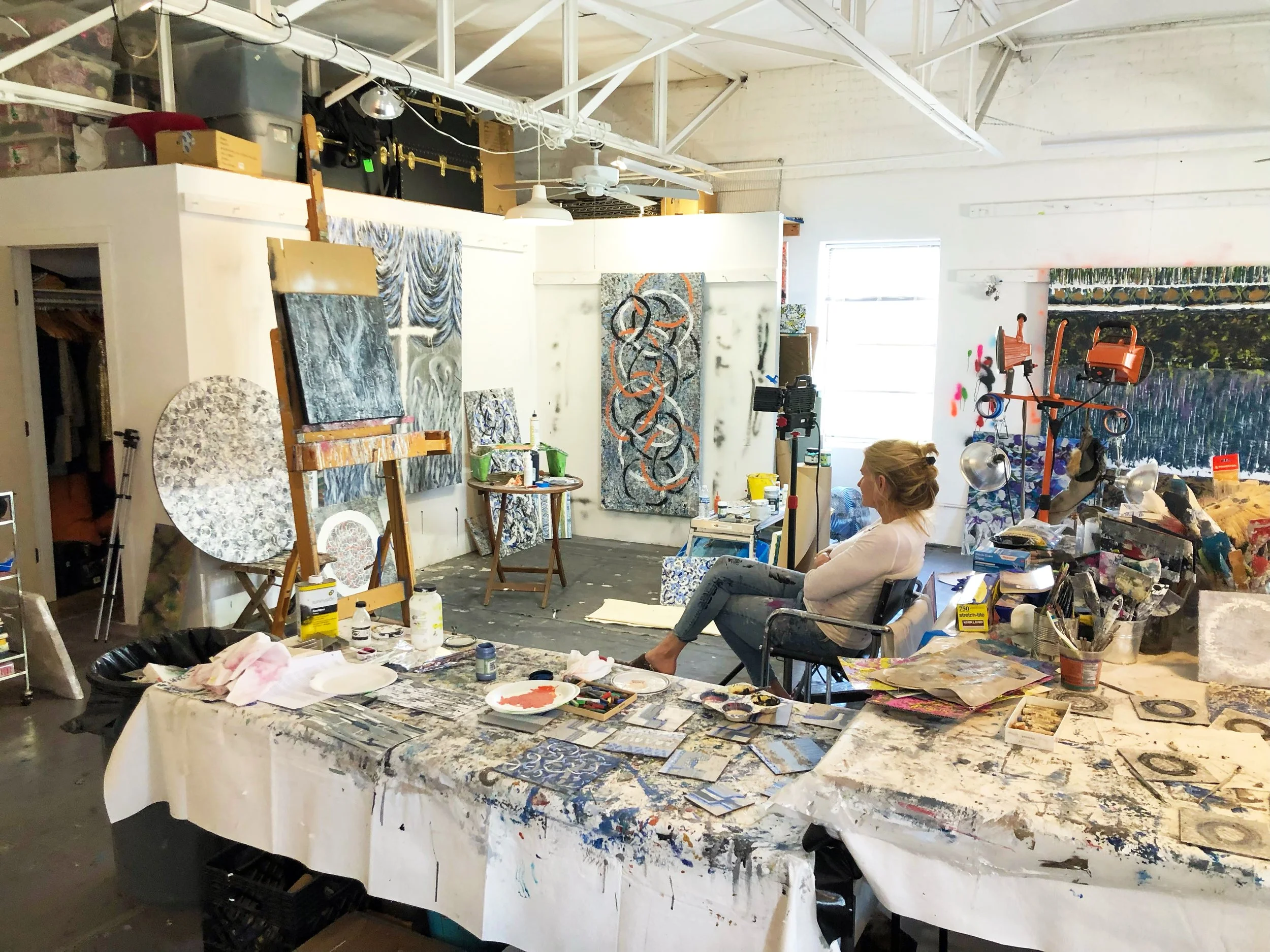 An artist's studio with paintings on the wall, a table covered with art supplies, and a woman sitting in a chair.
