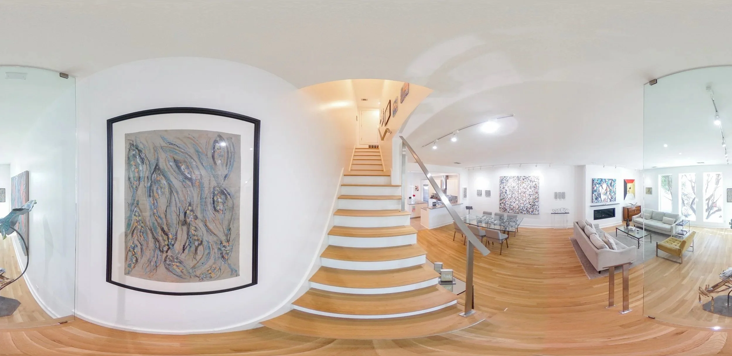 Interior of an art gallery or home with abstract paintings on white walls, hardwood floors, a staircase, a sitting area with sofas, chairs, and a fireplace, illuminated by ceiling lights.