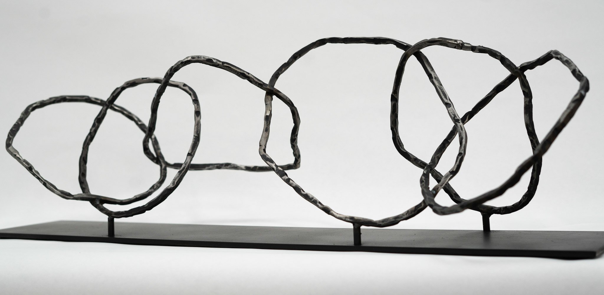 Abstract black metal sculpture with interconnected loops on a black base against a plain white background.