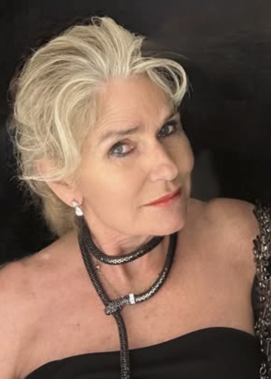 A woman with short, styled blonde hair lying down on a dark surface, wearing jewelry including earrings and layered necklaces, dressed in a strapless black top or dress.