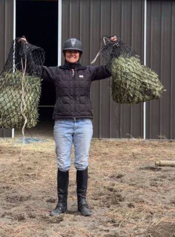 A person wearing a helmet, black jacket, and boots, standing outdoors, holding two large bags of what looks like plants or foliage.