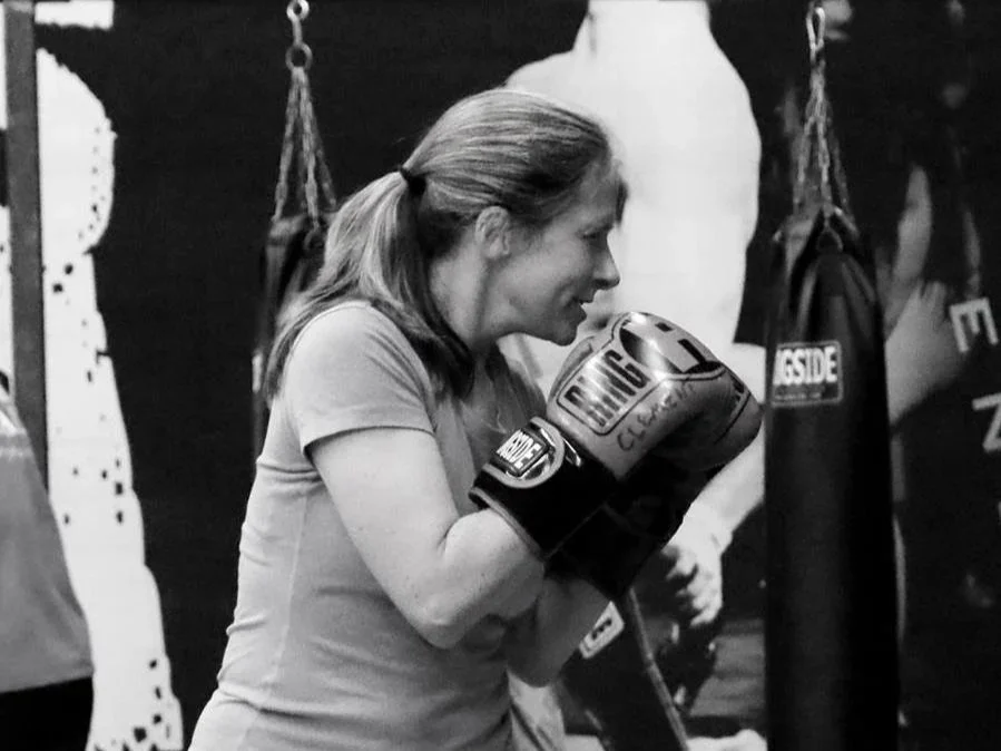 A woman with hair tied back in a ponytail practicing boxing, wearing boxing gloves and leaning forward in a fighting stance at a gym.