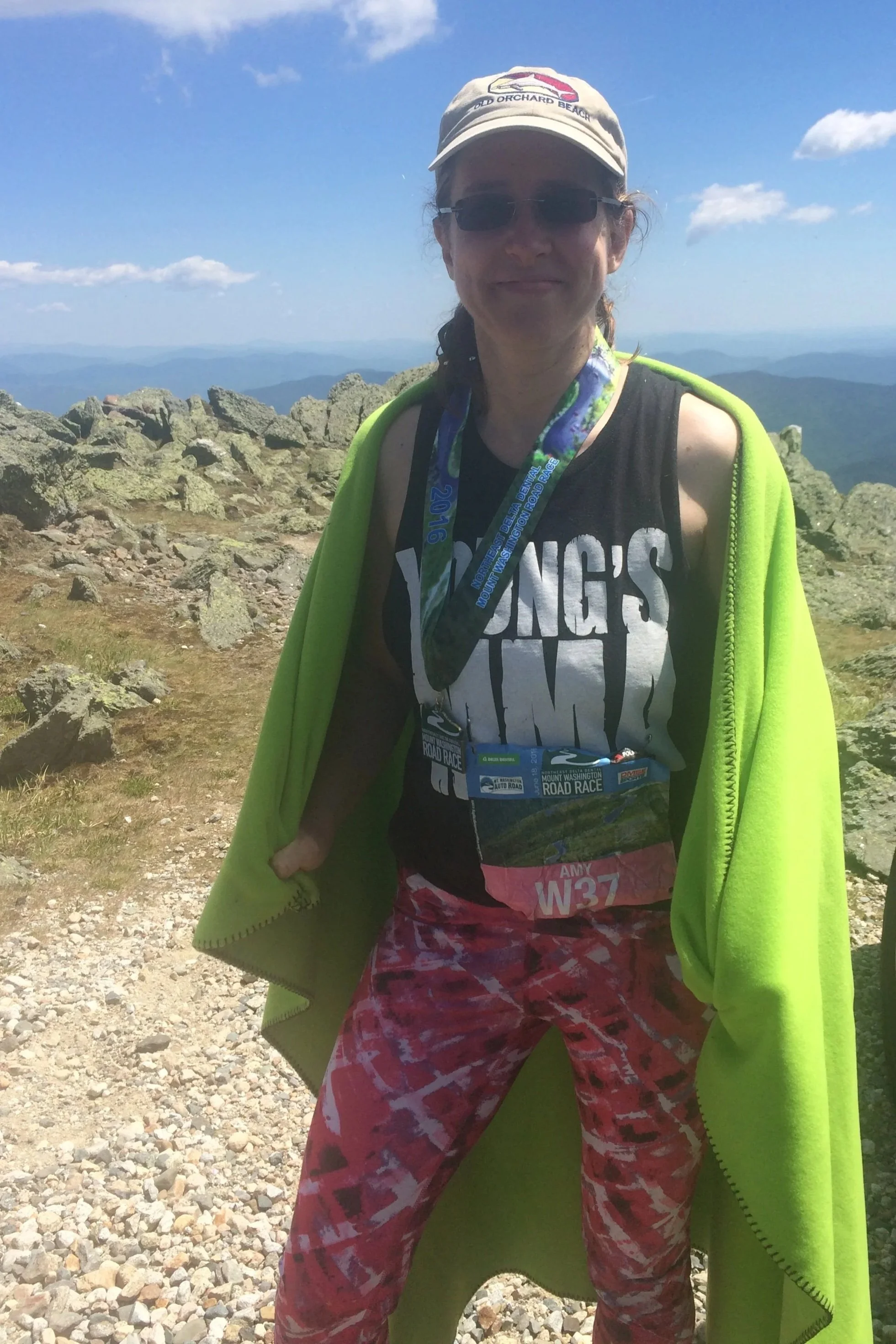 A woman wearing a beige cap, sunglasses, a black t-shirt, and red patterned pants stands on rocky mountain terrain with mountains and a blue sky in the background. She has a lime green jacket draped over her shoulders and a medal around her neck.