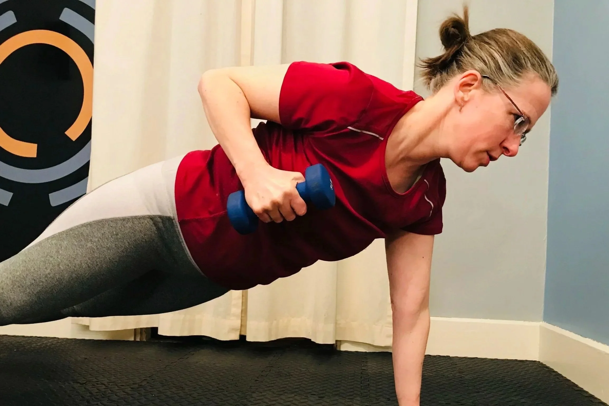 A woman in red workout clothes doing a plank exercise while holding a blue dumbbell in her right hand.