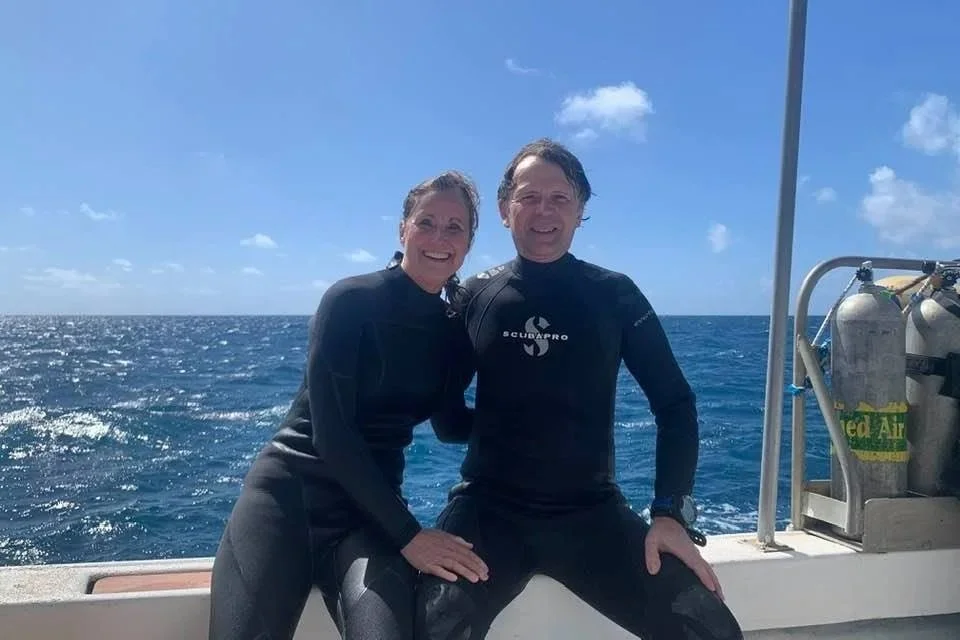 Two people smiling and posing together on a boat at sea, dressed in wetsuits, with the ocean and blue sky in the background.
