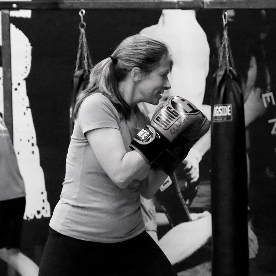 Woman practicing boxing in a gym, wearing boxing gloves and in a fighting stance near punching bags.