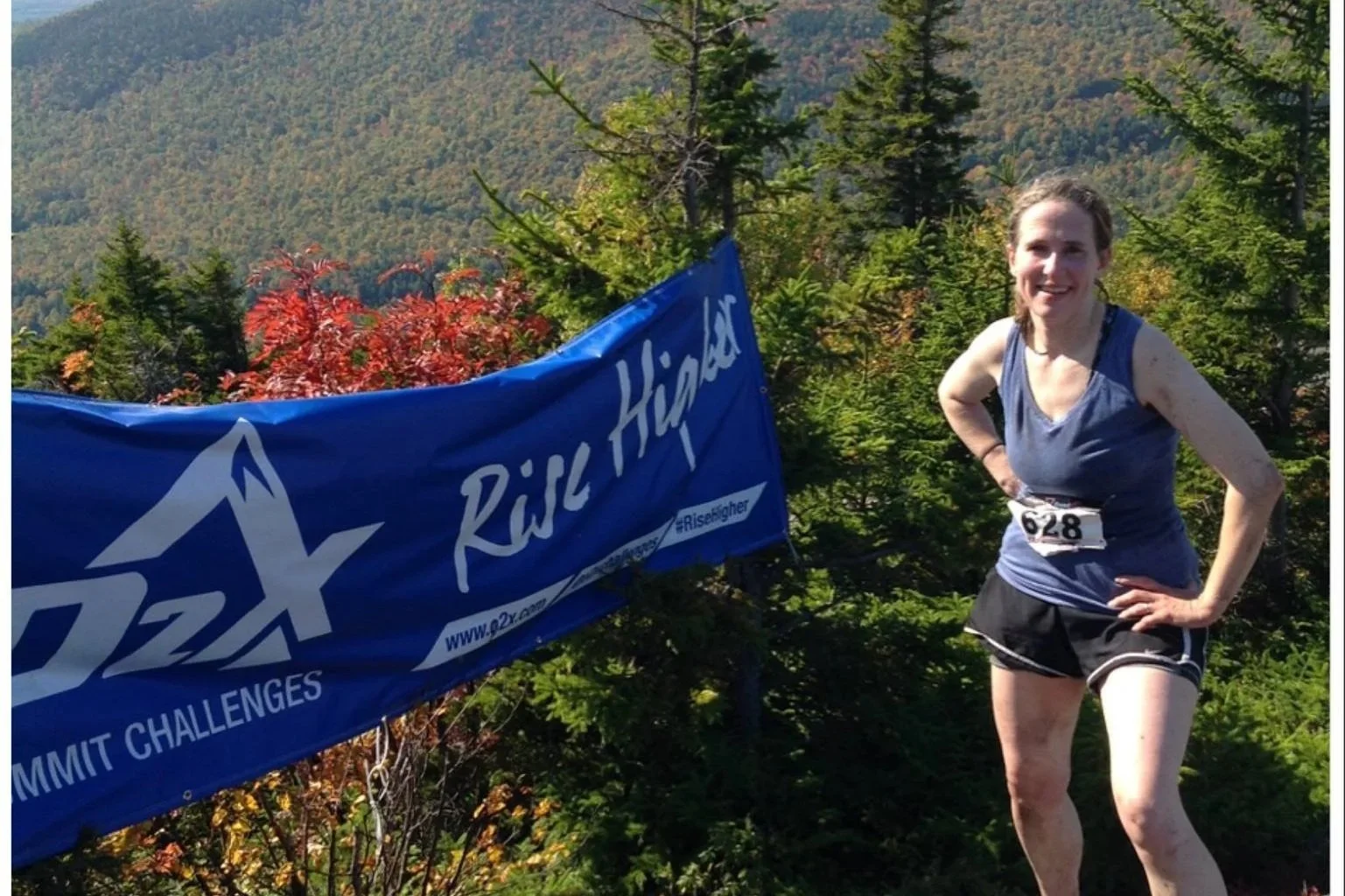 A woman with race number 628 on her clothes, dressed in a blue tank top and black shorts, smiling and standing outdoors in a forested mountain area with green and red trees, next to a blue banner that reads 'Q2X RID High
